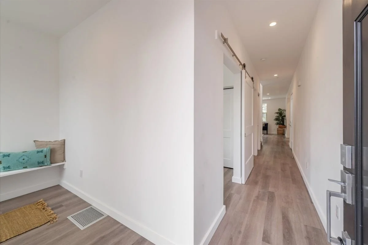 Empty hallway in a modern home with light-colored walls, wooden flooring, and recessed ceiling lights. A glimpse of a room with a plant and furniture at the end of the hall.