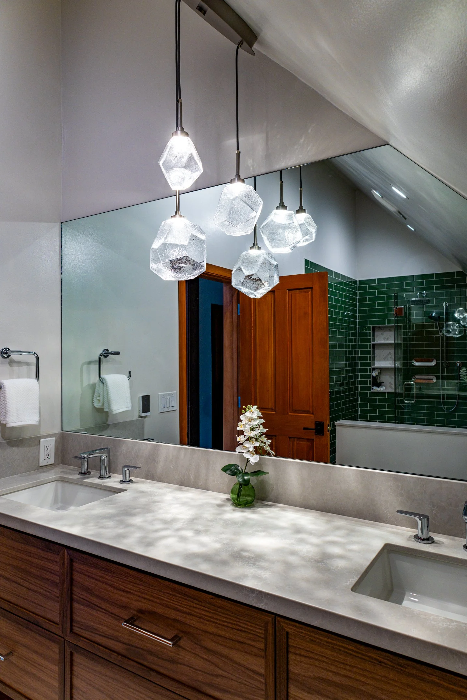 A bathroom with a large mirror reflecting a vanity with a beige stone countertop, two sinks, and a small green vase with white flowers. Overhead, a cluster of six geometric pendant lights hangs from the ceiling. In the reflection, a wooden door and a