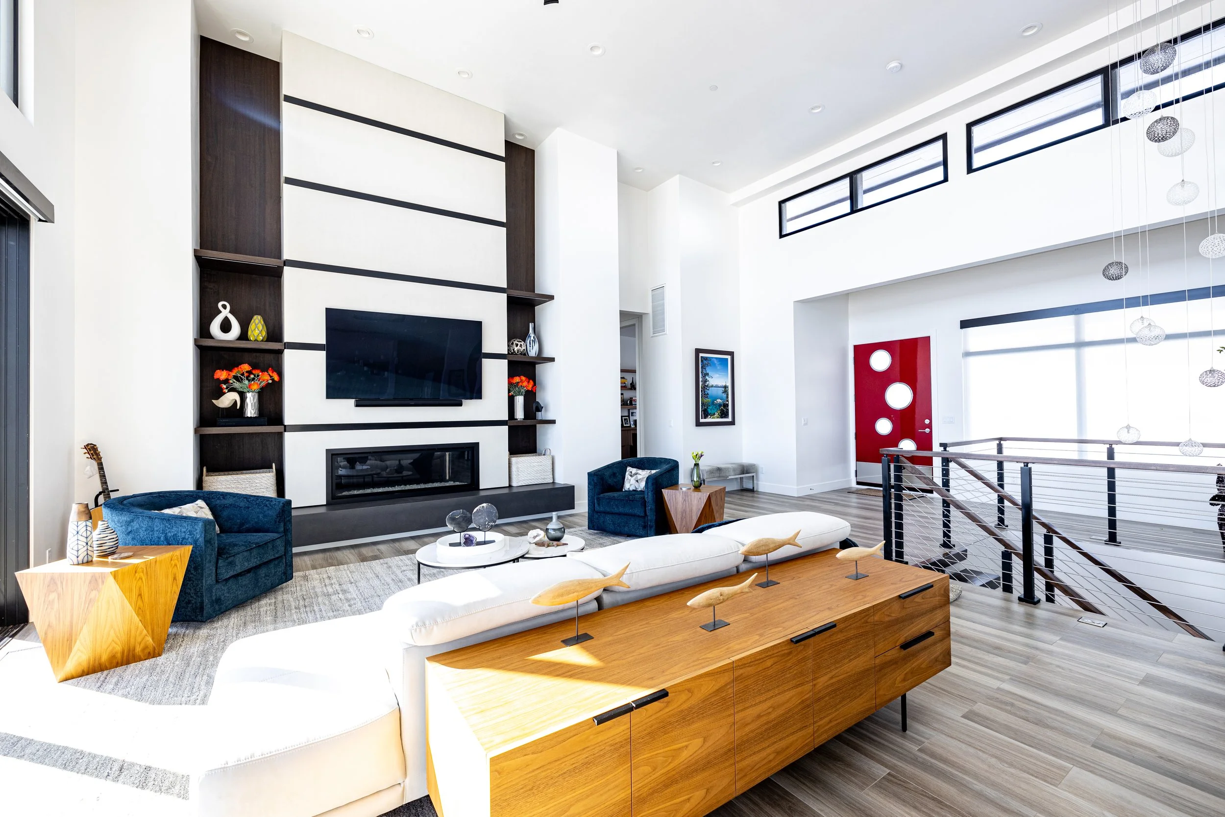 Modern living room with white walls, high ceiling, and large windows. Features a fireplace, mounted TV, blue armchairs, a cream-colored sofa with decorative fish sculptures, and wooden furniture. Red front door with circular windows and a staircase w
