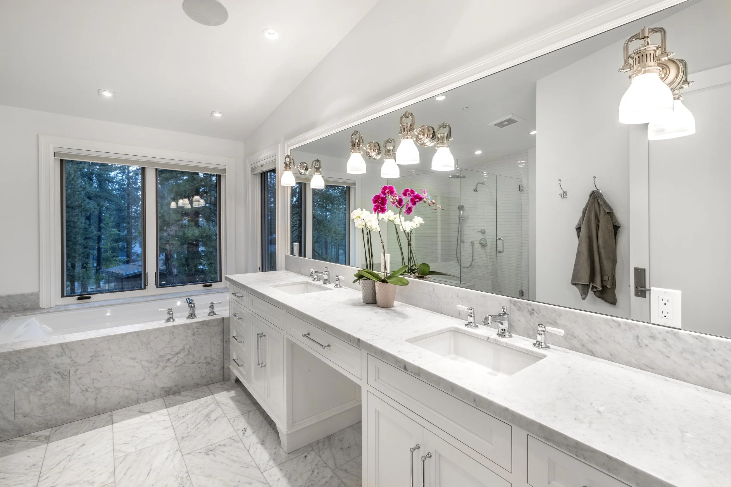 Bright white bathroom with marble countertops, double sinks, a large mirror, orchids, a bathtub near a window, and a glass shower enclosure.