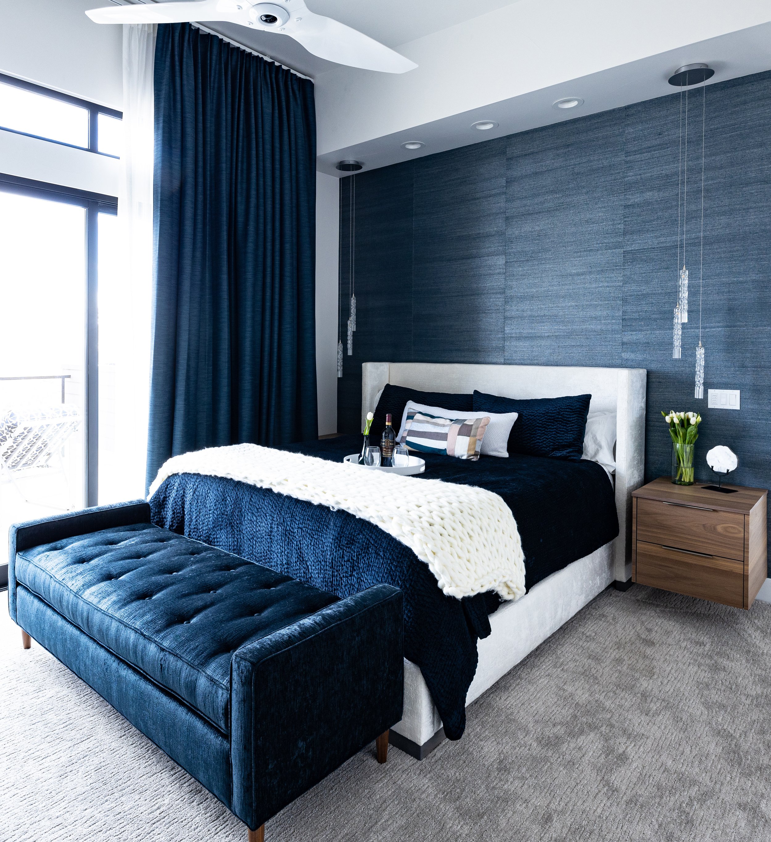 Modern bedroom with blue textured accent wall, large window with dark blue curtains, white bed with black and patterned pillows, black bedspread, white chunky-knit throw blanket, black tufted bench at the foot of the bed, wooden nightstand with flowe