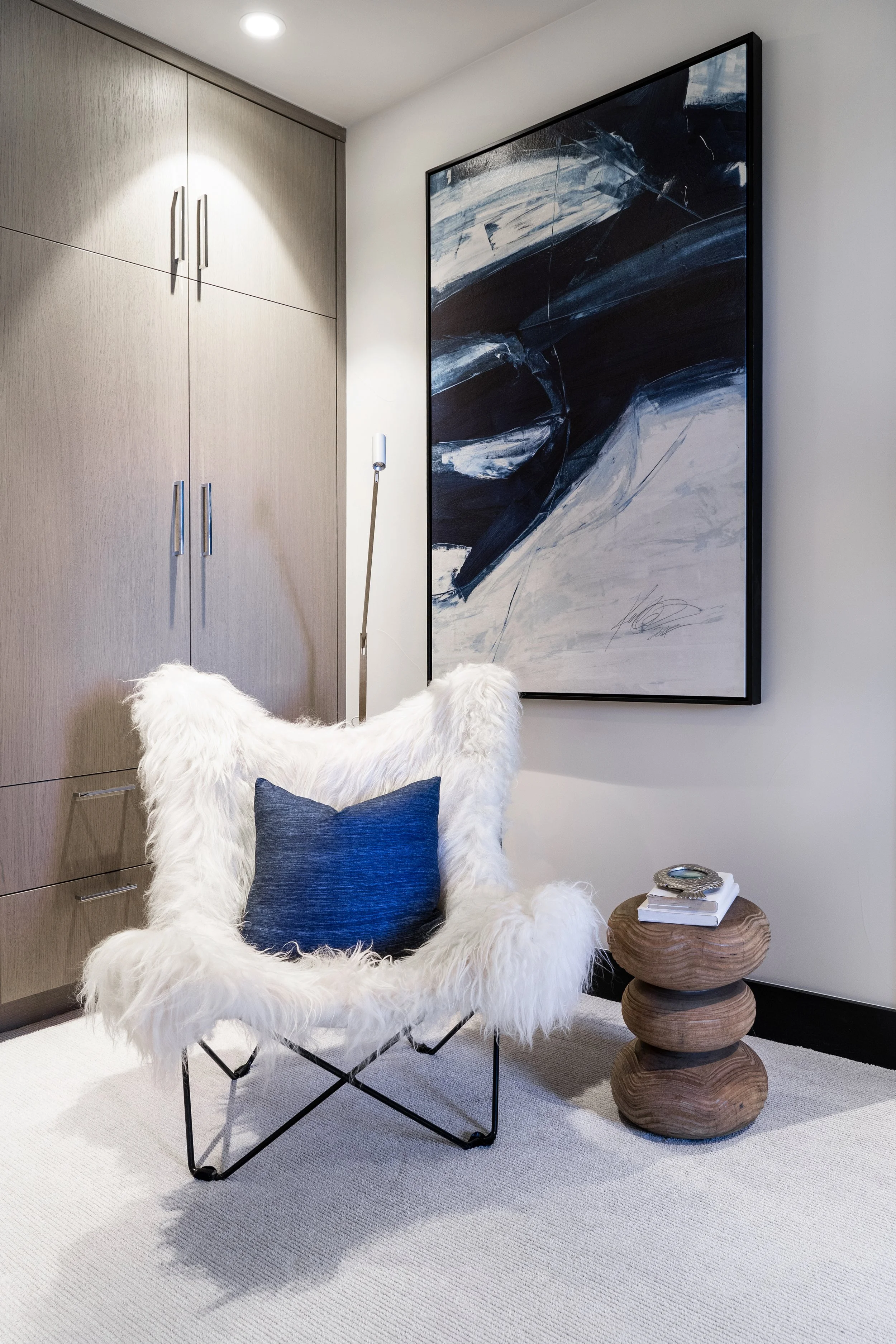 Modern interior corner with a white fluffy chair, a dark blue cushion, a wooden side table with books and jewelry, a large abstract black and white painting, a light-colored wooden cabinet, and a tall floor lamp.
