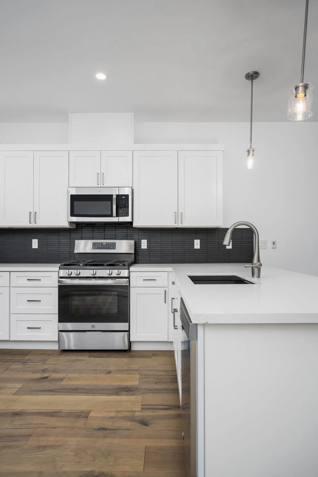 Modern kitchen with white cabinets, stainless steel appliances including a microwave, oven, and dishwasher, black backsplash, and wooden flooring.