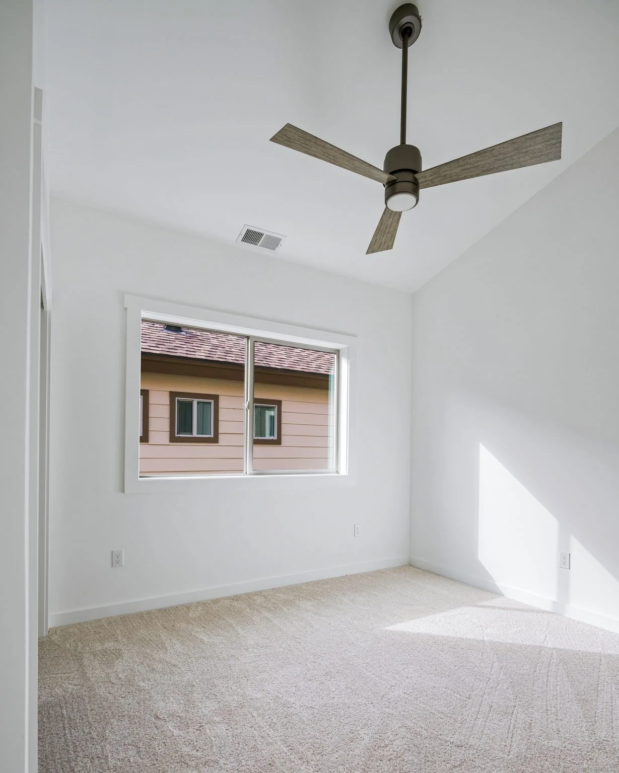 Empty room with white walls, beige carpet, a window showing neighboring house, ceiling fan, and electrical outlets.