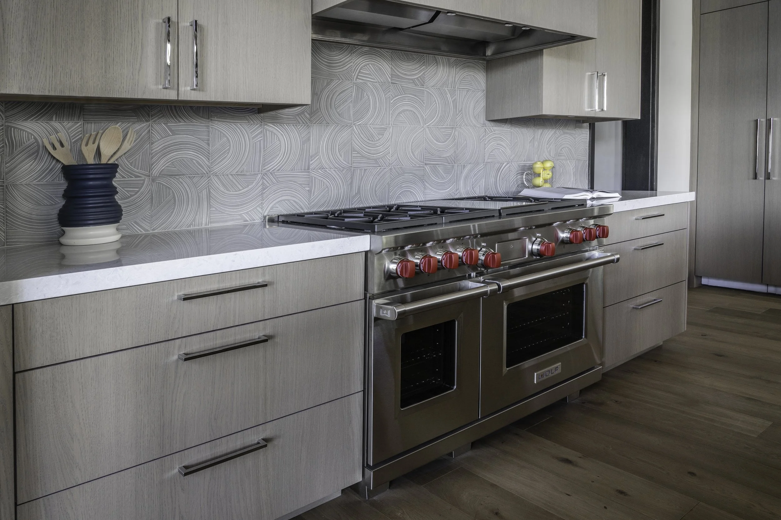 Modern kitchen with a Wolf range with red knobs, light wood flat panel cabinets, Kelly Wearstler organic patterned tile backsplash, and European Oak wood flooring.