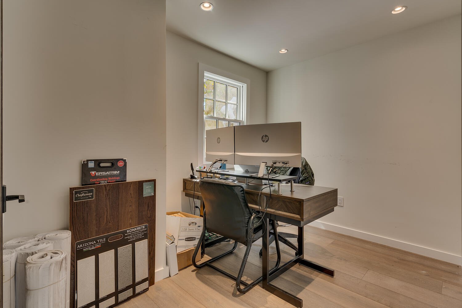 An office space with a wooden desk, two HP monitors, a black leather office chair, and window with natural light. Building materials and tile samples are leaning against the wall.