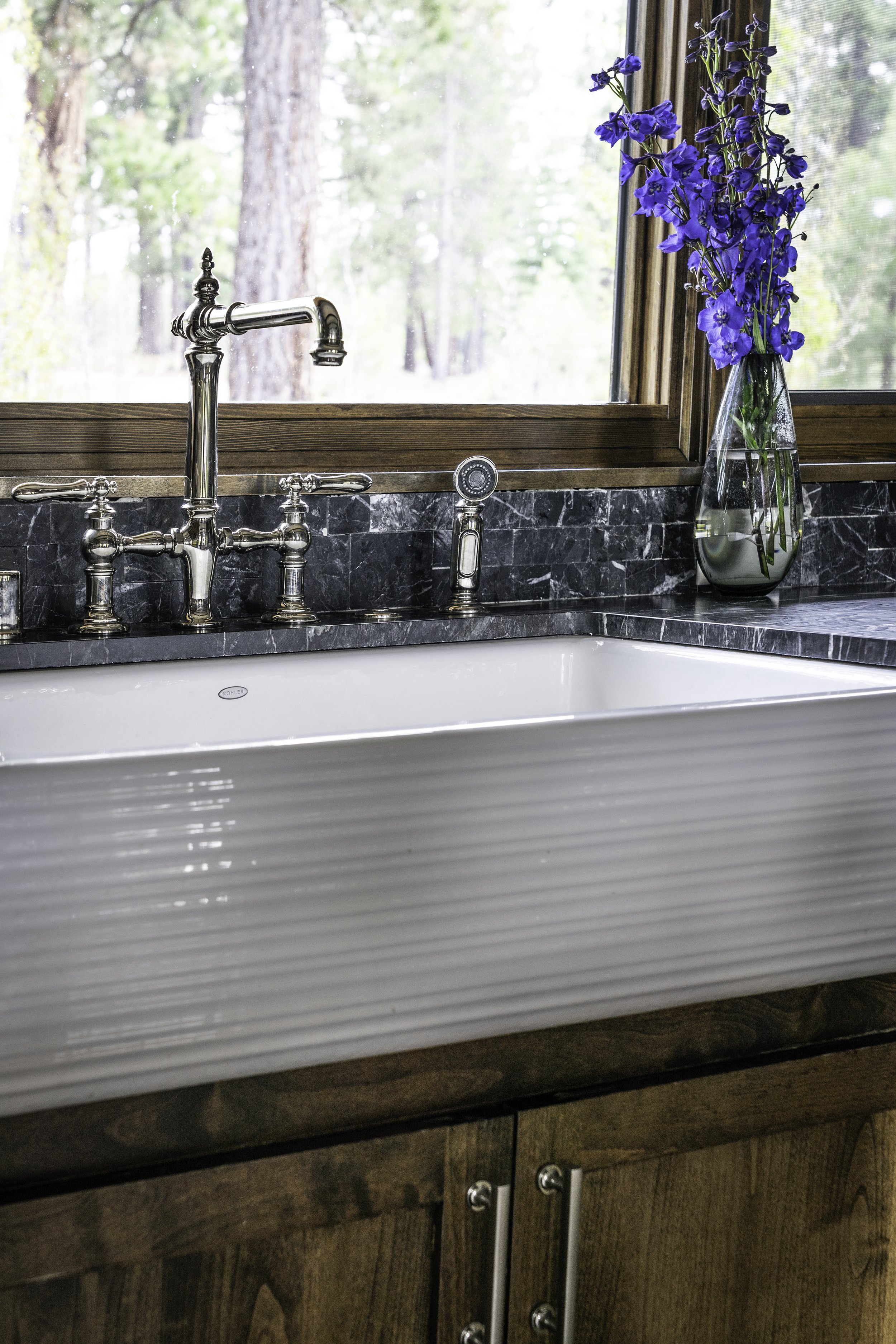 A farmhouse-style kitchen sink with a dark marble countertop, a vintage-style silver faucet, a bottle of soap or sanitizer, and a tall glass vase with blue delphinium flowers in front of a window showing trees.
