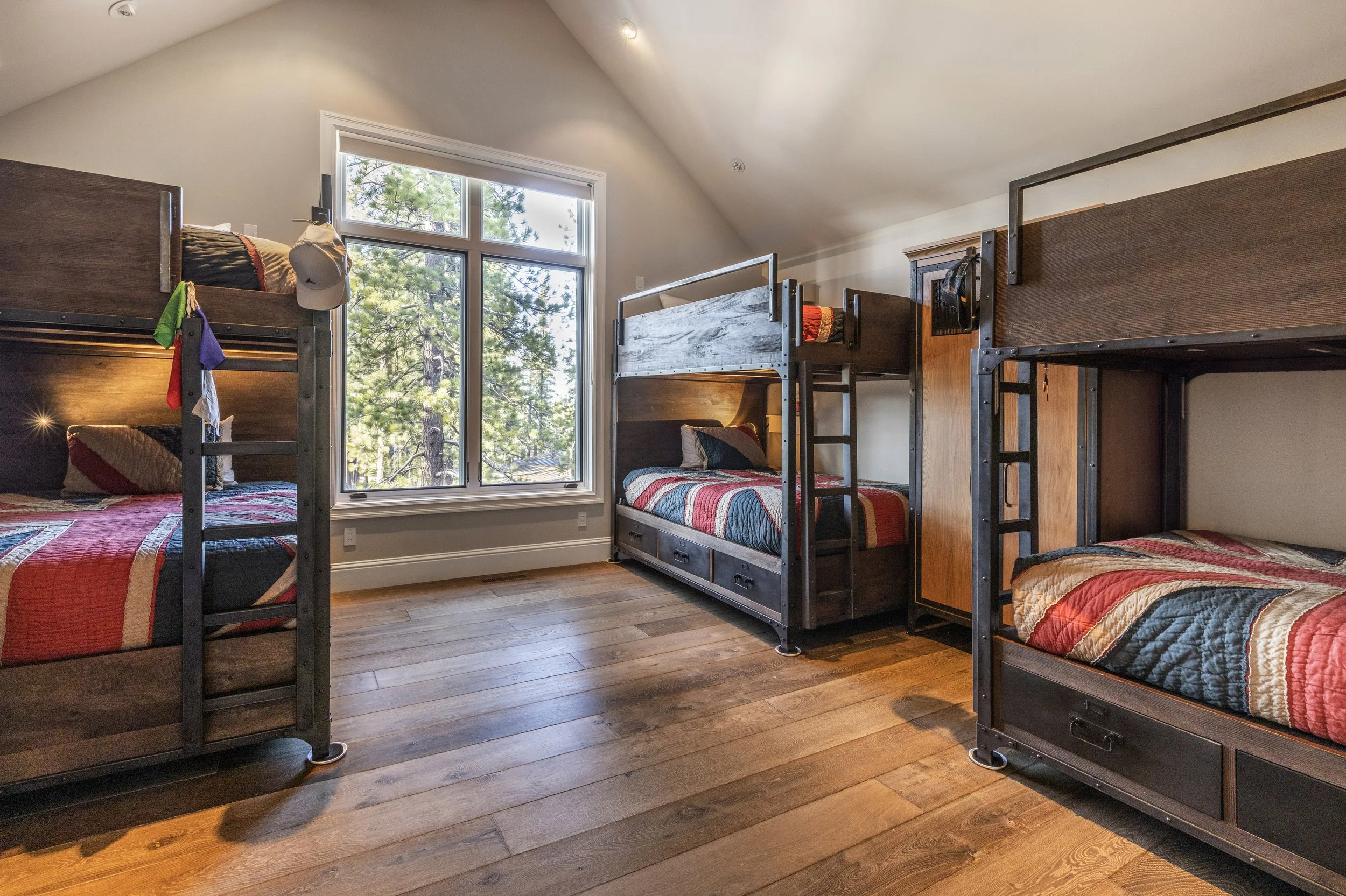 A bedroom with three sets of bunk beds, each with a quilt featuring a red, black, and beige pattern. There is a large window letting in natural light, showing tall pine trees outside. The room has wooden floors and neutral-colored walls.