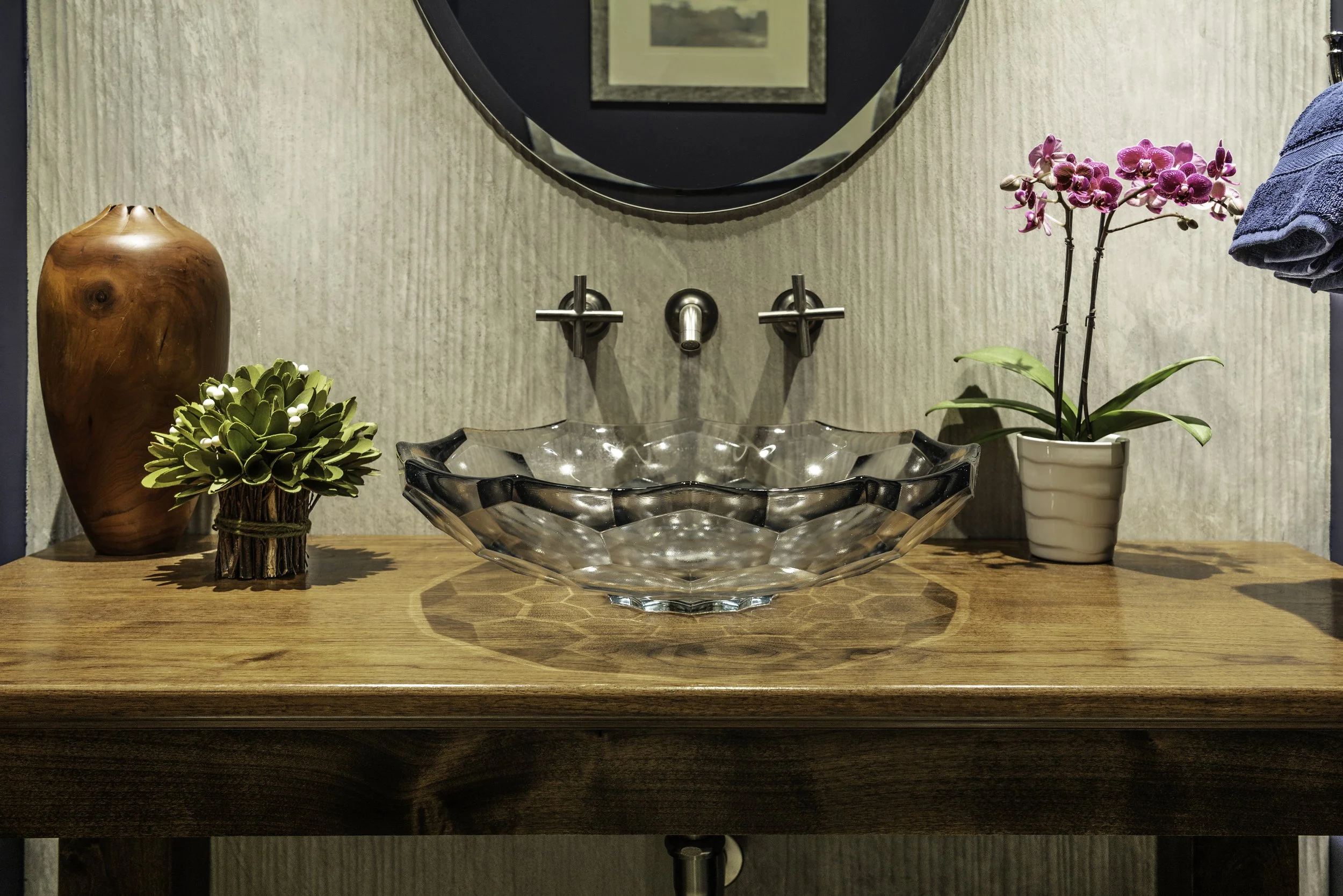 A wooden bathroom vanity with a glass vessel sink. Plants and decorative items are placed on the vanity, with a round mirror and framed picture above it.