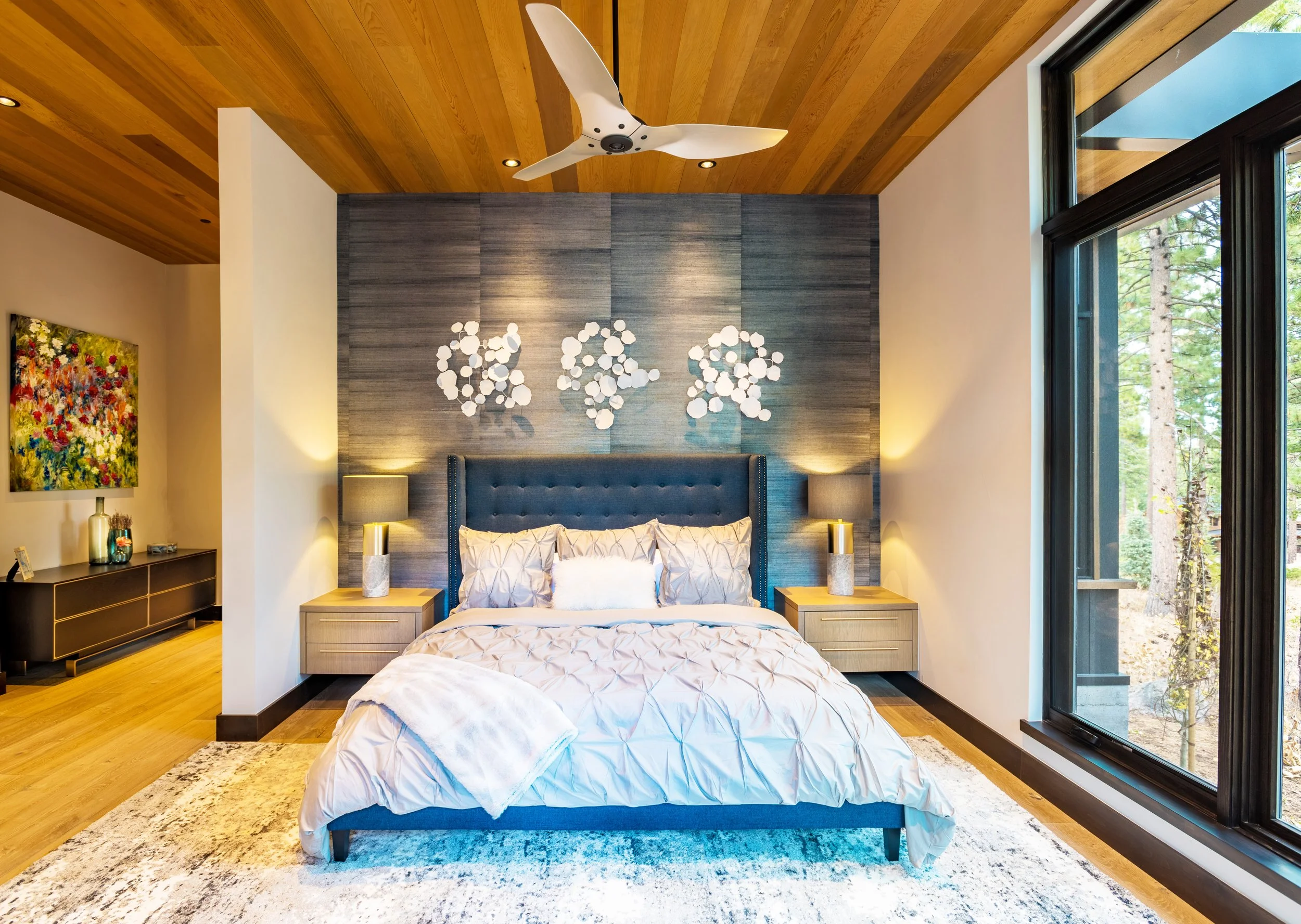 Modern bedroom with a large bed, gray upholstered headboard, white bedding, and decorative pillows. Two nightstands with matching lamps flank the bed. A dark accent wall with decorative wall art is behind the bed. Large windows to the right let in na