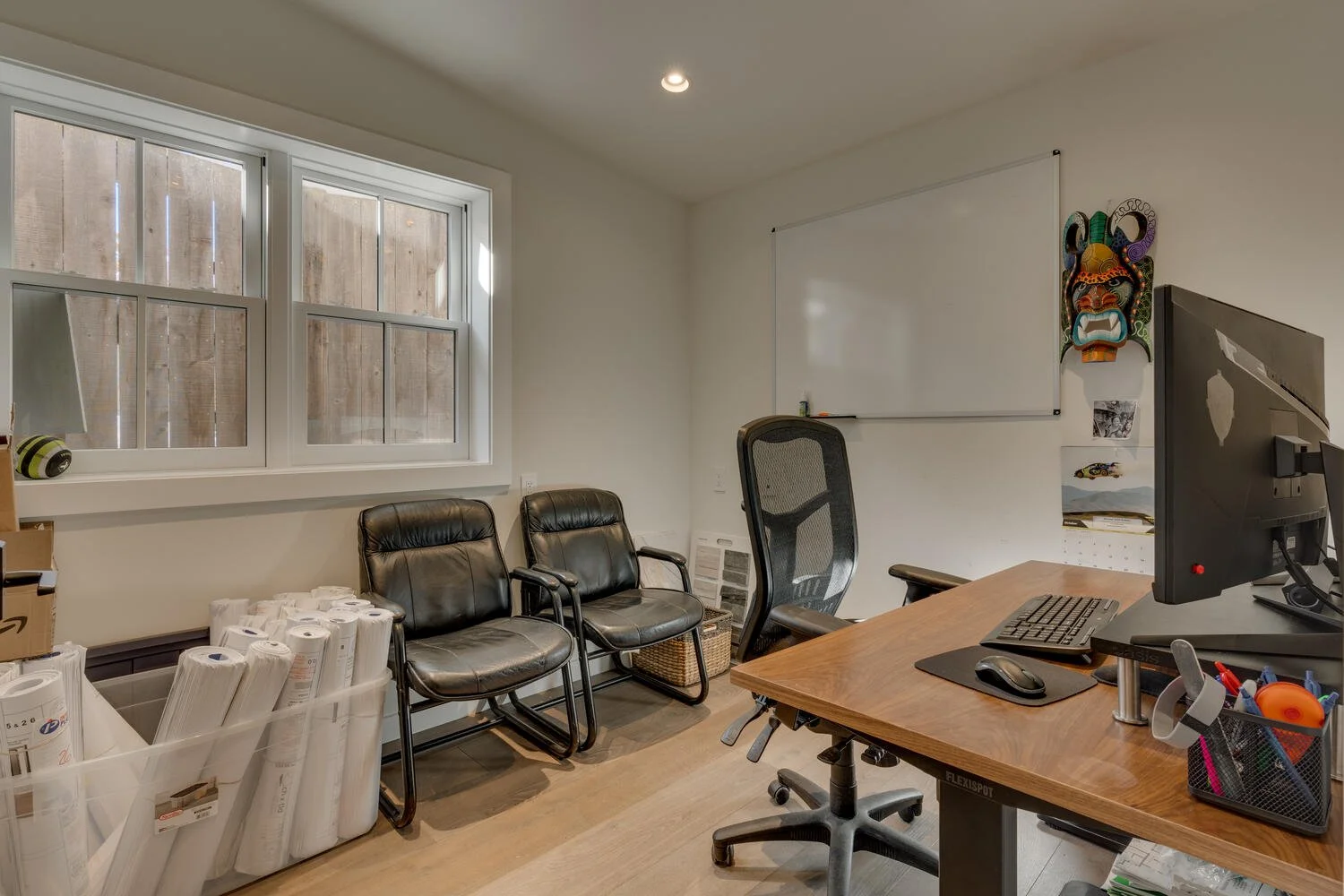 An office space with a wooden desk, computer, and office chair. There are two black leather chairs, a whiteboard, and various office supplies, including rolls of paper and a desk organizer. A window with wooden blinds is on the left side, and a color