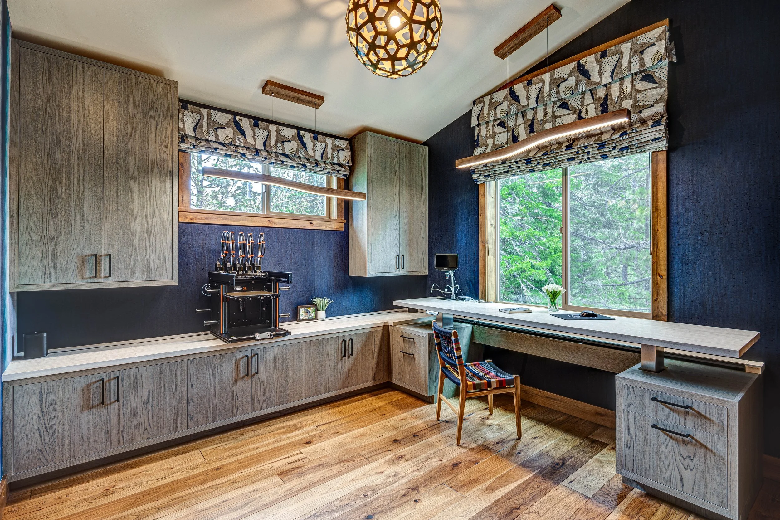 Home office with wooden cabinets and desk, a 3D printer, large windows with patterned curtains, and lush greenery outside. Modern lighting fixtures illuminate the space.