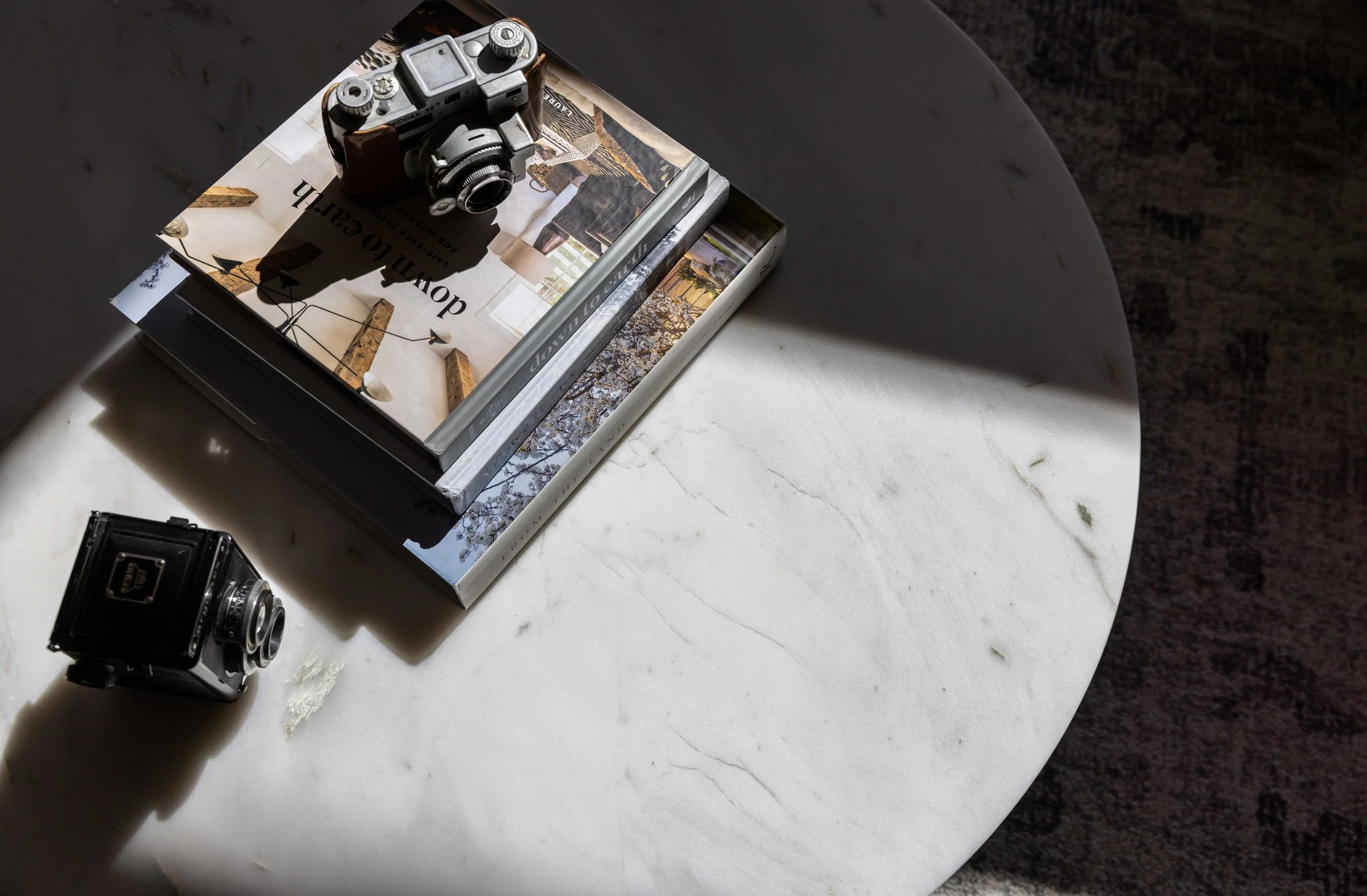 A white marble round table with sunlight casting shadows on it, holding a stack of magazines, a vintage camera, and a small black camera.