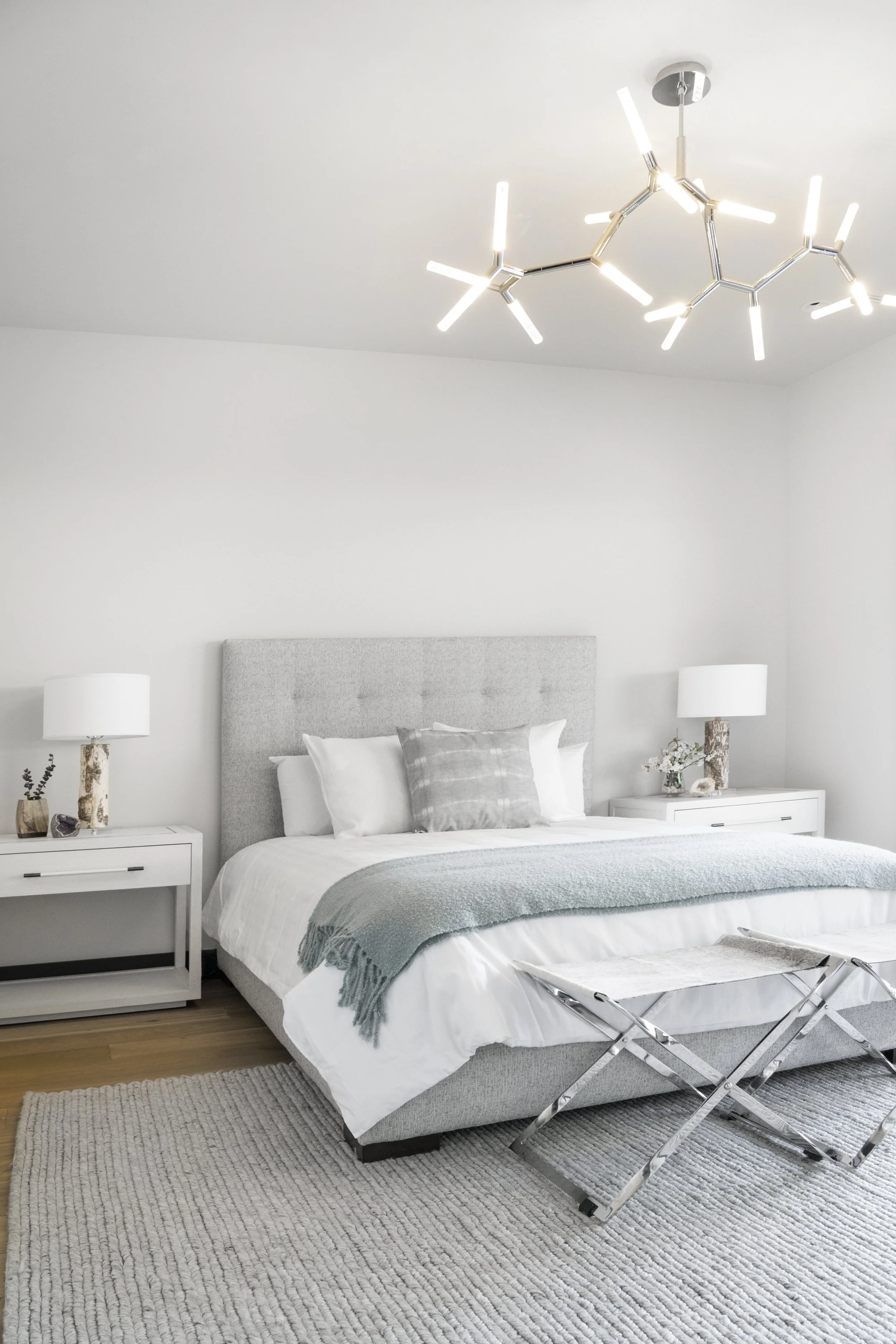 Modern bedroom with gray upholstered bed, white bedding, gray blanket, two white nightstands with lamps, and a contemporary ceiling chandelier, with a cream textured rug on wooden floor.