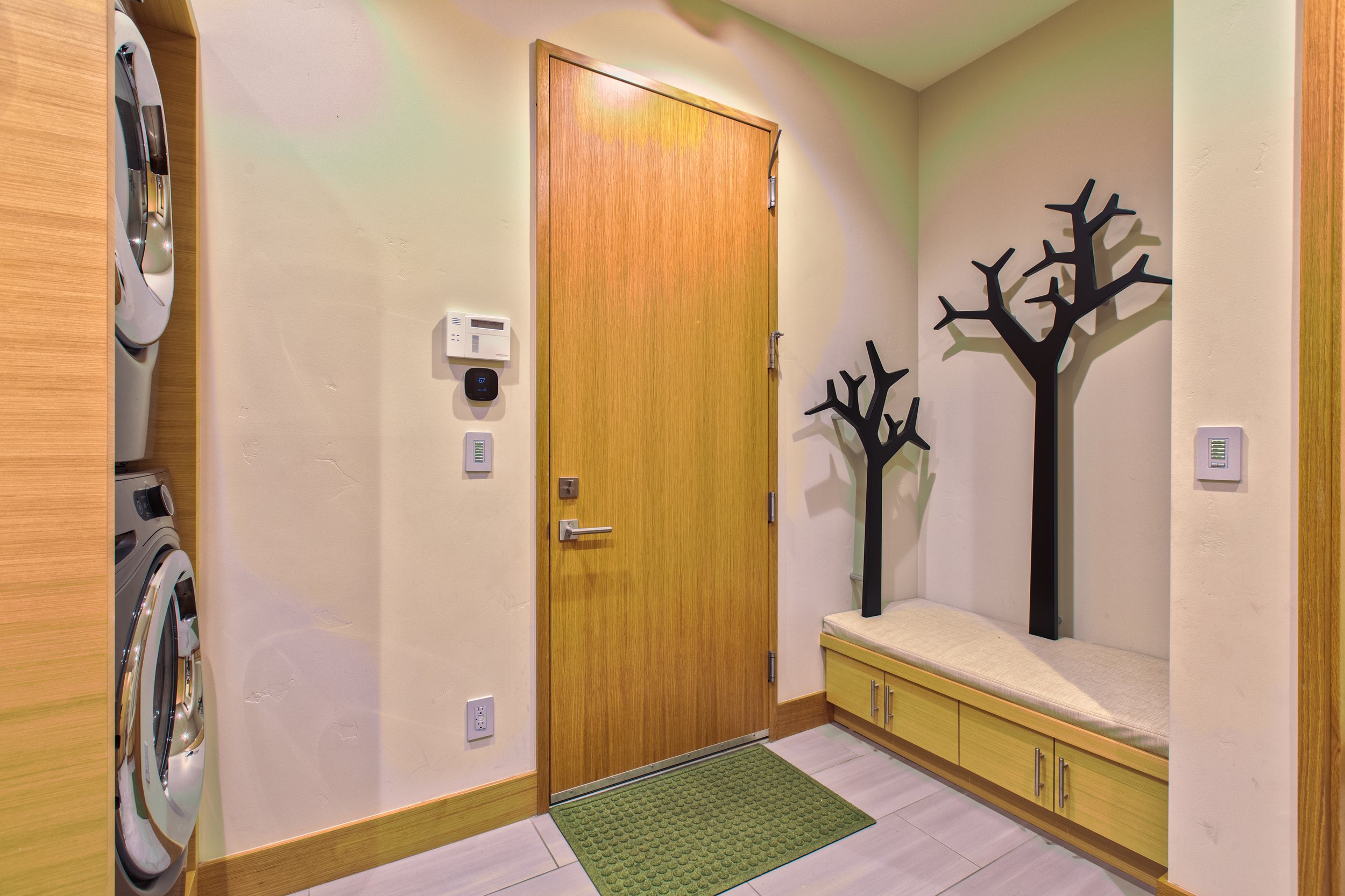 Entryway with wooden door, wall-mounted thermostat, electrical switches, a green doormat, and a bench with two black tree-shaped coat racks.