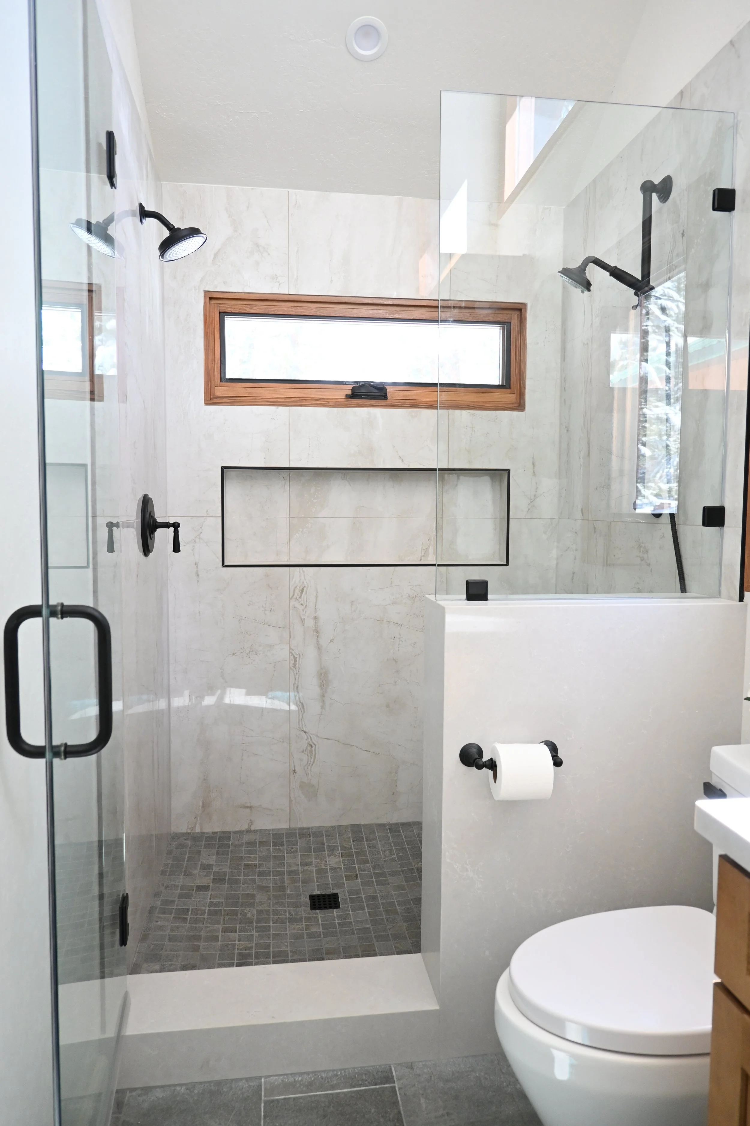 Modern bathroom with walk-in shower, black fixtures, and a toilet, featuring a small horizontal window and minimalist decor.
