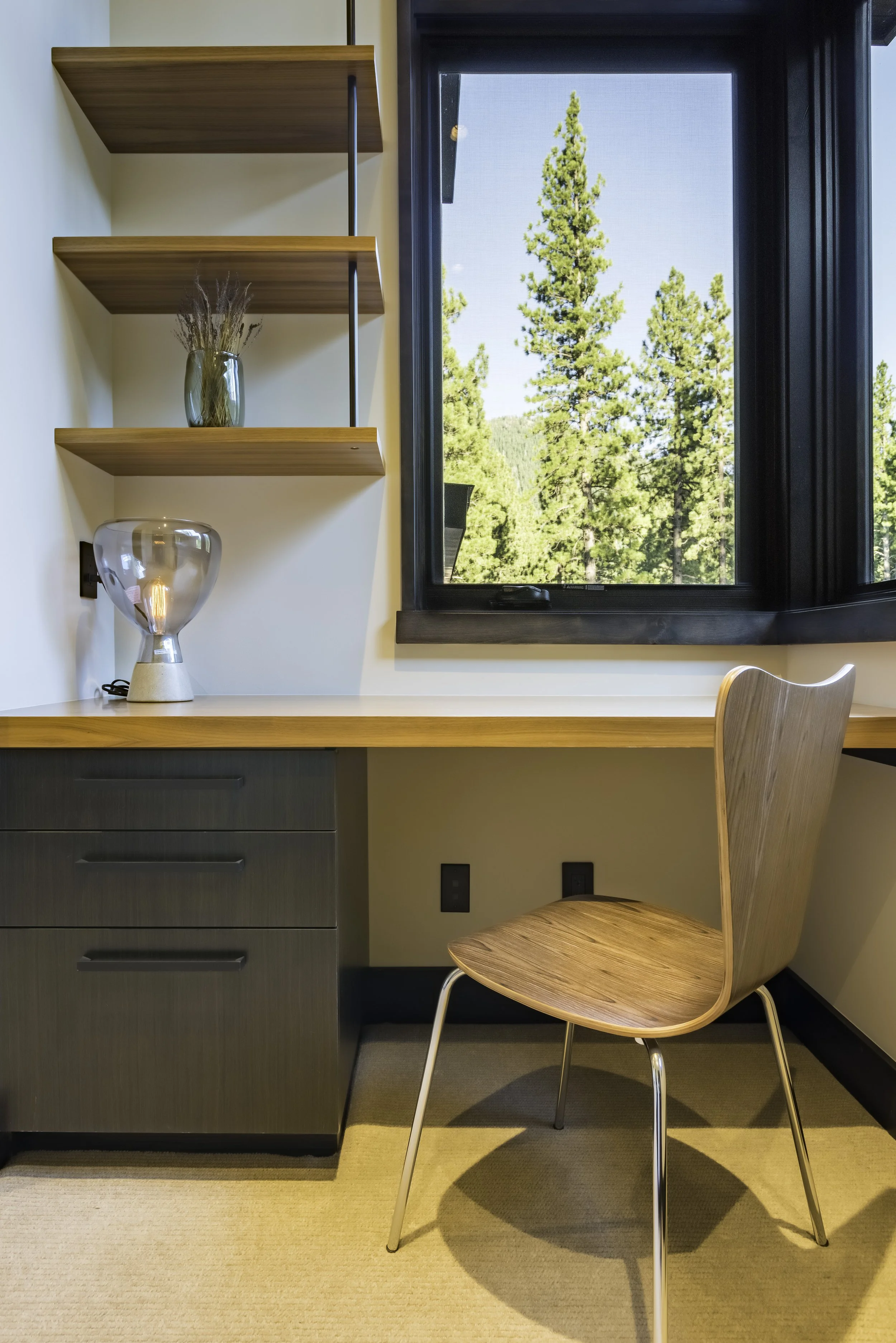A minimalist workspace with a wooden desk, a wooden chair, and floating shelves with a vase and decorative object, near a window overlooking trees.