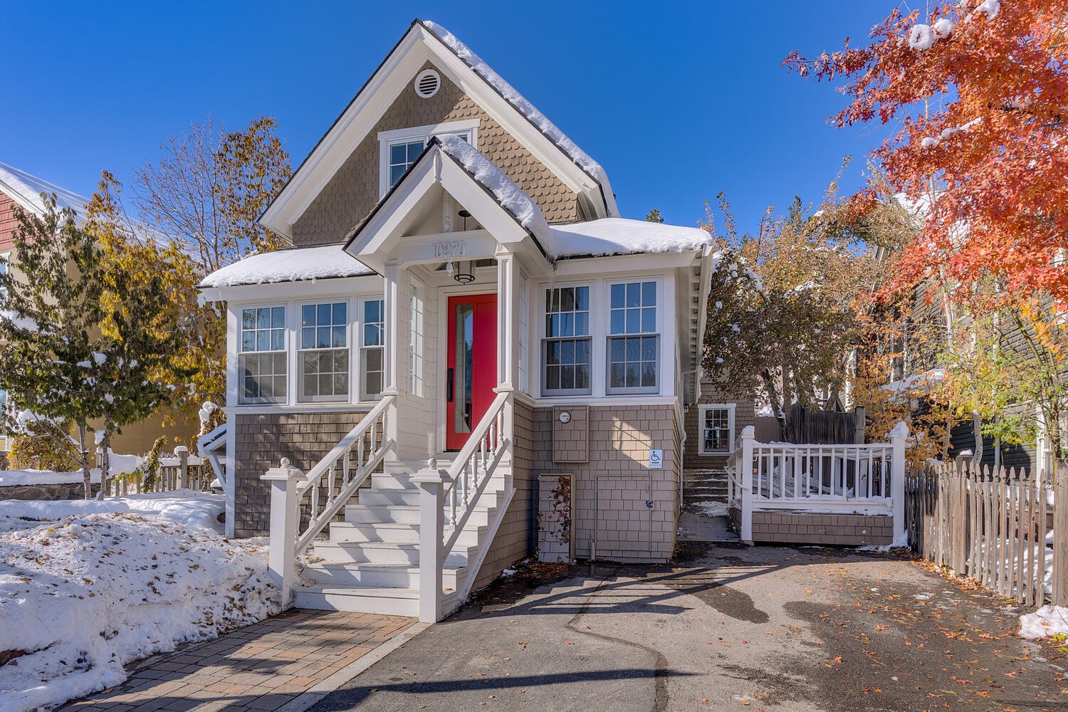 Front of a two-story house with a red door, white stairs and railing, surrounded by snow and autumn-colored trees, under a clear blue sky.
