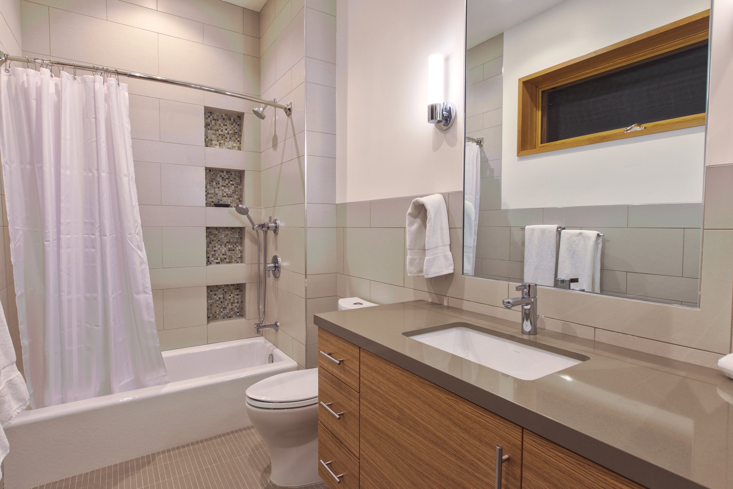 Modern bathroom with bathtub, shower, toilet, and wooden vanity with sink, large mirror, white towels, and small window.