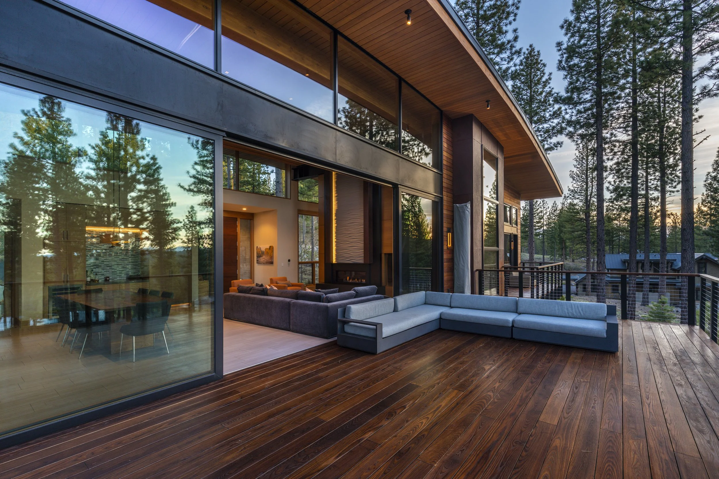 Modern house exterior with large glass windows and a spacious wooden deck surrounded by tall pine trees at sunset.