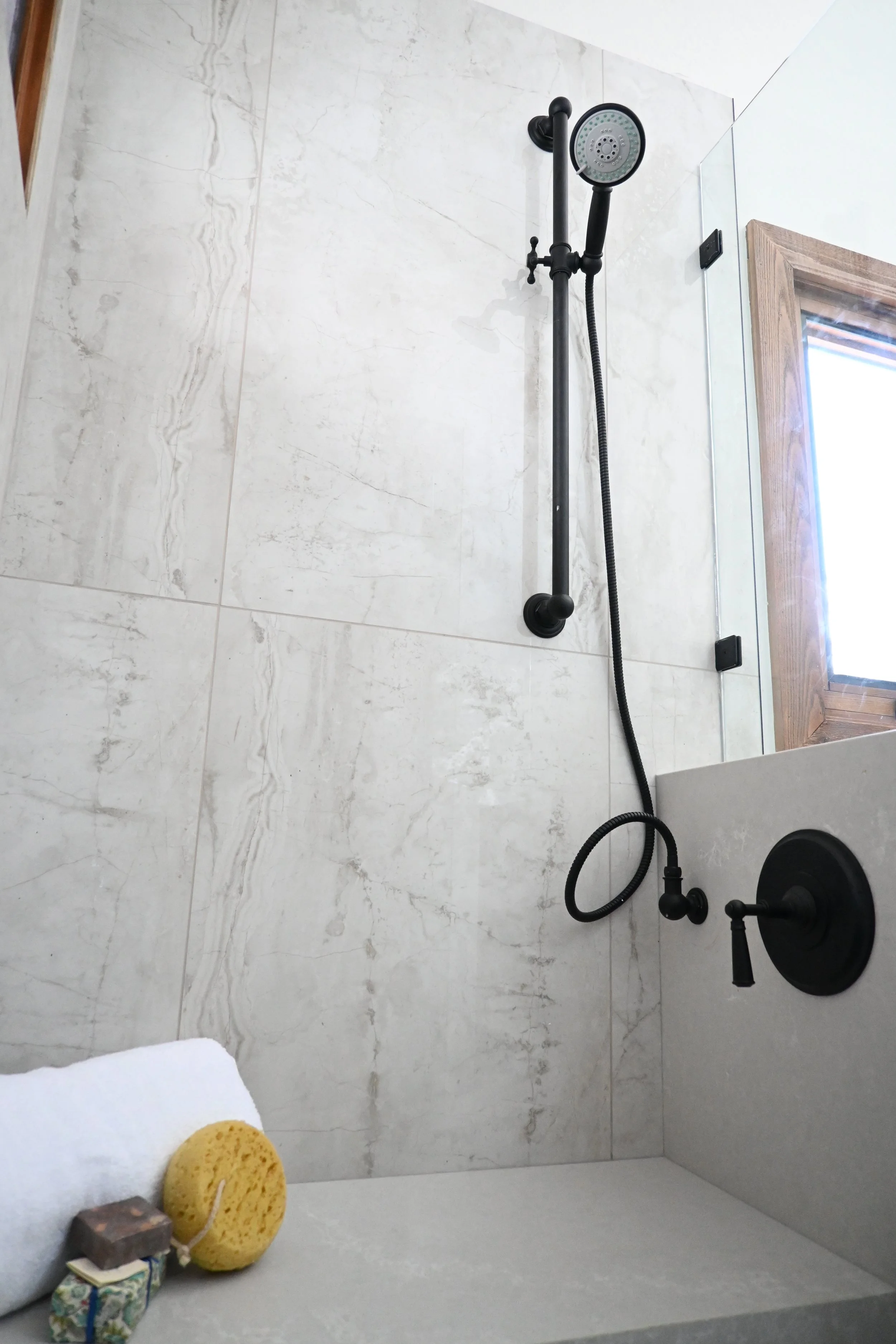 Shower with black fixtures and a handheld shower head, tiled wall, small window, and bath accessories including a sponge, a soap, a rolled towel, and a small box.