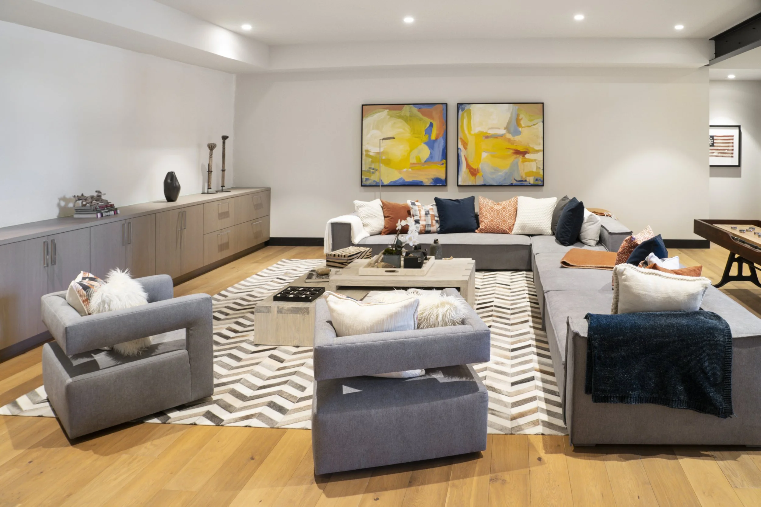 A modern living room with a large gray sectional sofa, a smaller gray armchair, and a patterned area rug. The sofa has multiple colorful pillows, and a low coffee table with decorative items. Abstract artwork hangs on the white wall behind the sofa, 