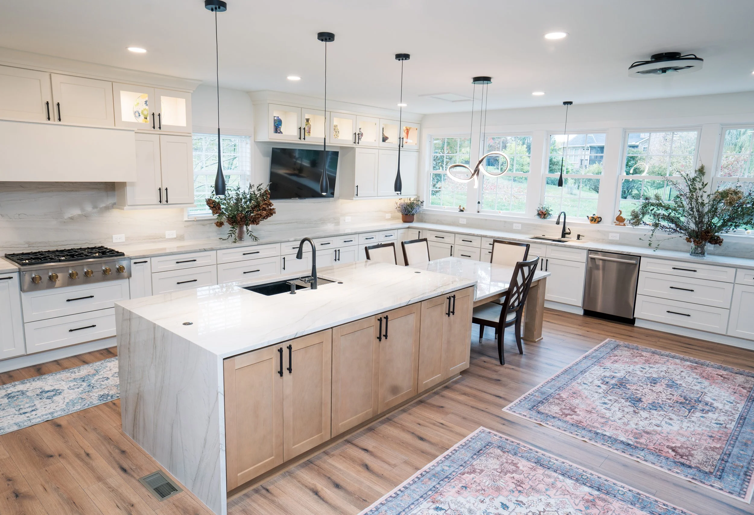 Modern kitchen with white cabinets, marble countertops, wooden island, and hardwood floors. Large windows provide natural light. Decor includes potted plants and a large vase with flowers. Dining table with chairs is adjacent to the window, and decor
