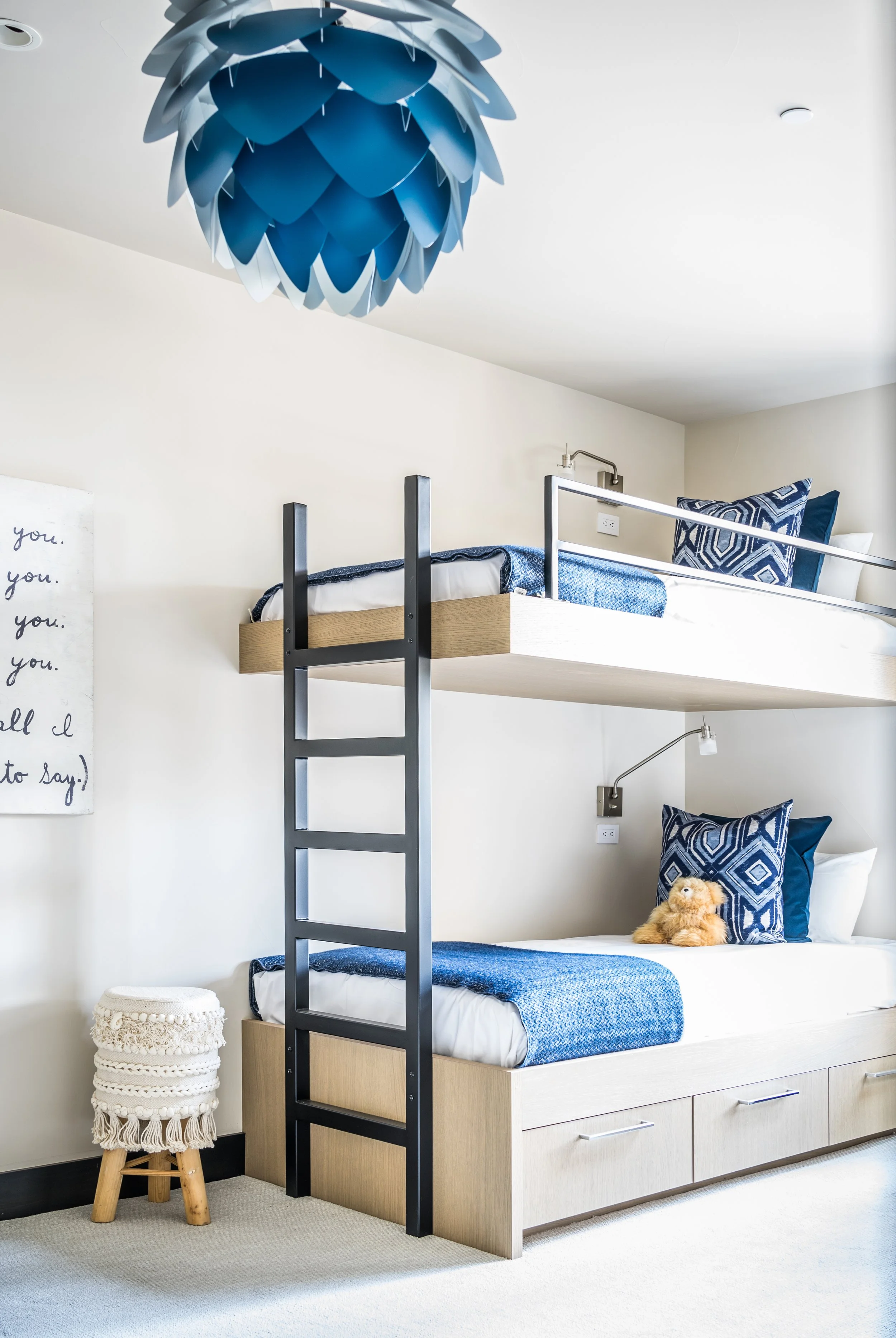 A bedroom with a bunk bed; the lower bed has a teddy bear and a blue geometric pillow, the upper bed is made with blue and white bedding, all against a white wall with a decorative quilted wall hanging.