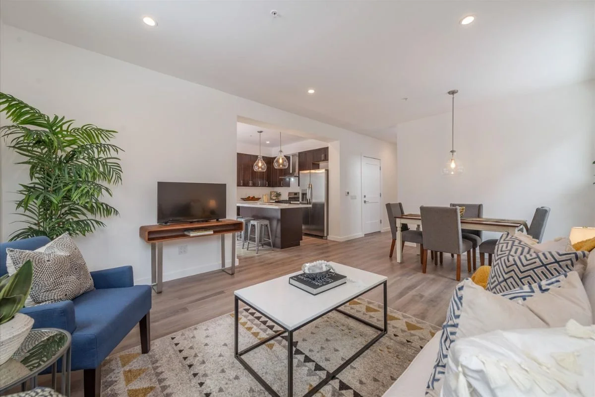 Open-concept living room and dining area with minimalistic decor, including a blue armchair, a white coffee table, a gray sofa with decorative pillows, a flat-screen TV on a wooden console, a dining table with six chairs, a hanging pendant light, and
