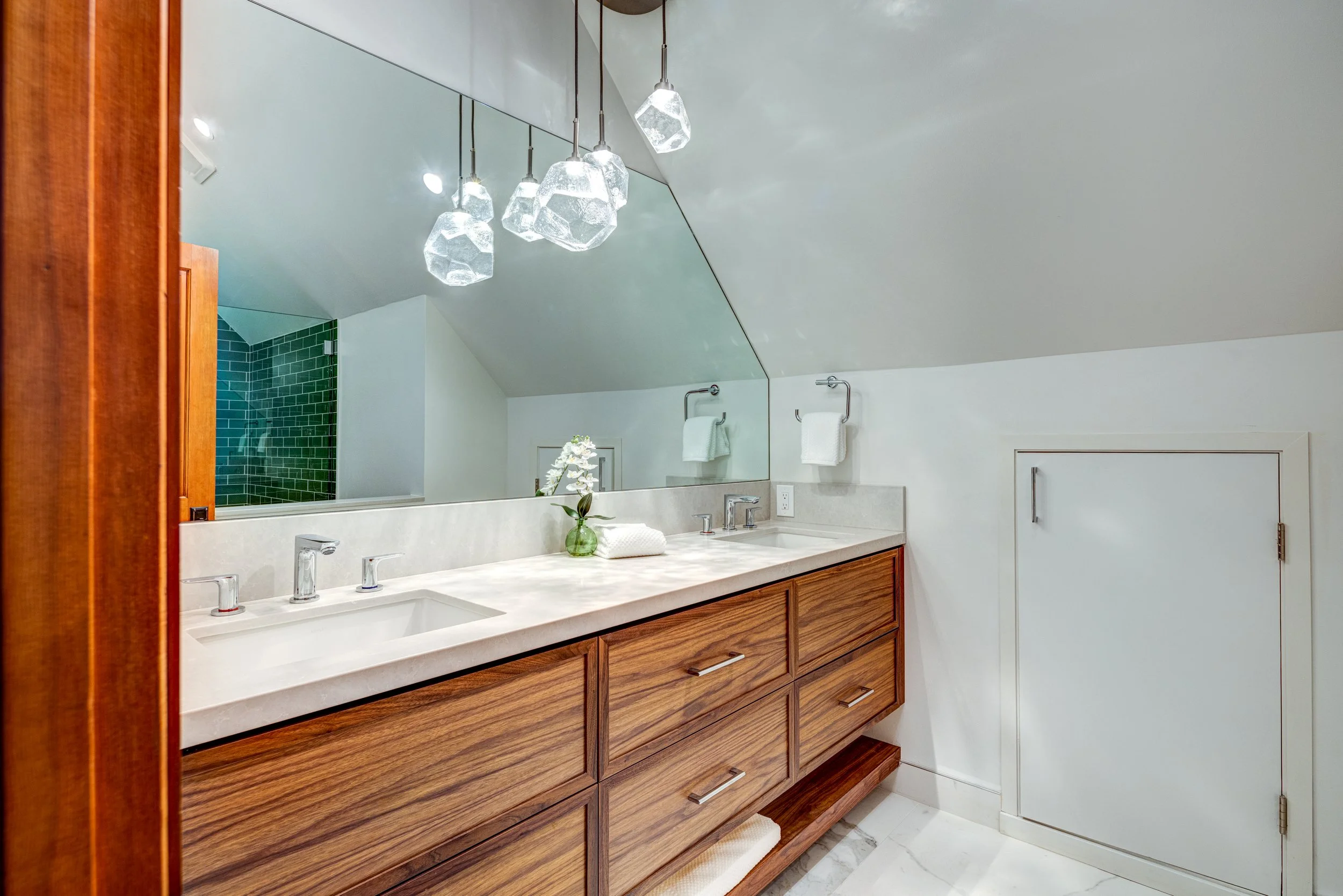 Bathroom vanity with a large mirror, two sinks, wooden drawers, a small green vase with white flowers, towels, modern hanging light fixtures, and a glimpse of a green-tiled shower area.