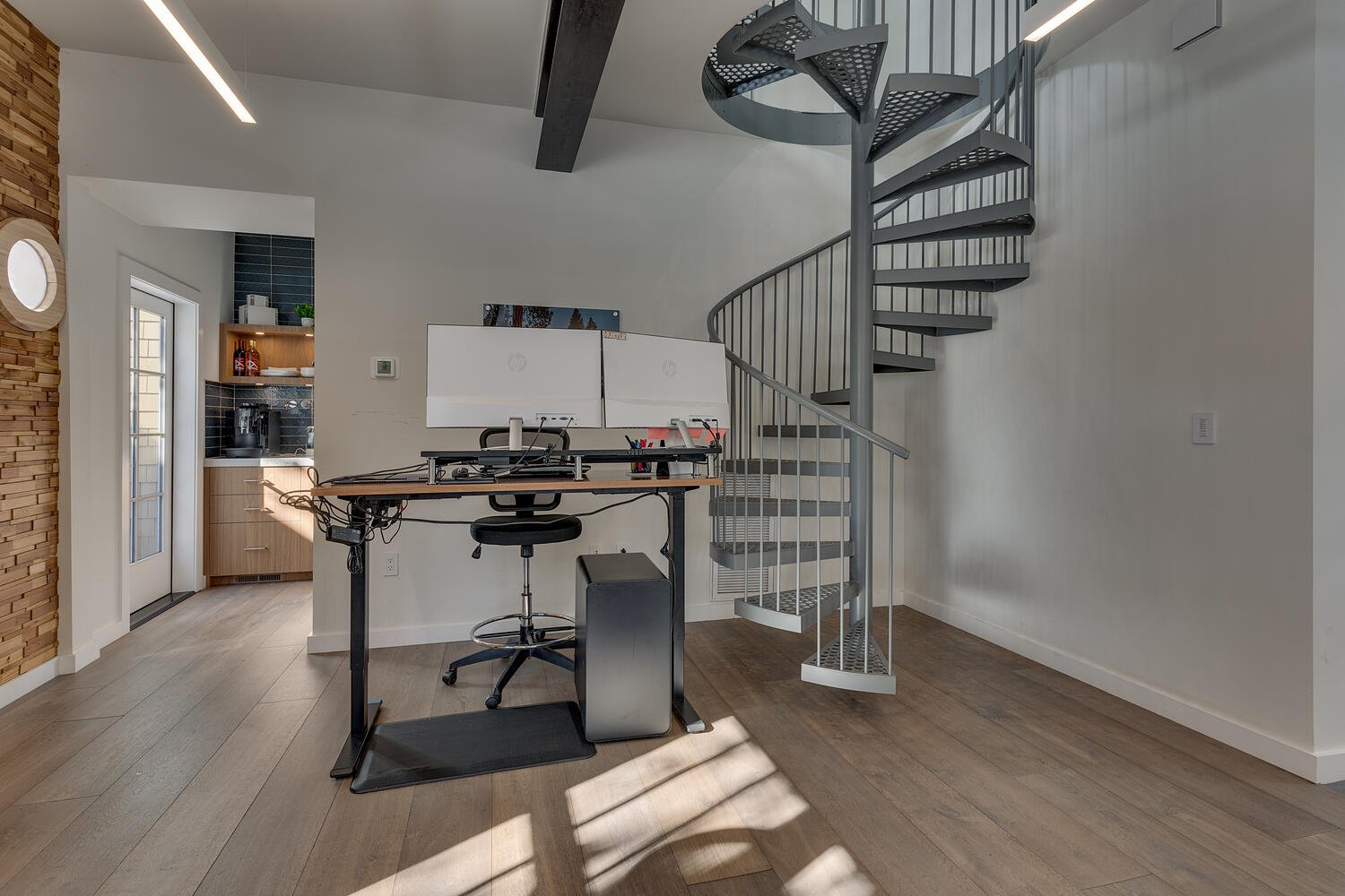Home office with a standing desk featuring two monitors, a black office chair, and a computer tower. Spiral staircase on the right leads upstairs. Kitchen area with dark backsplash and wooden cabinets in the background. Natural light streaming in cre