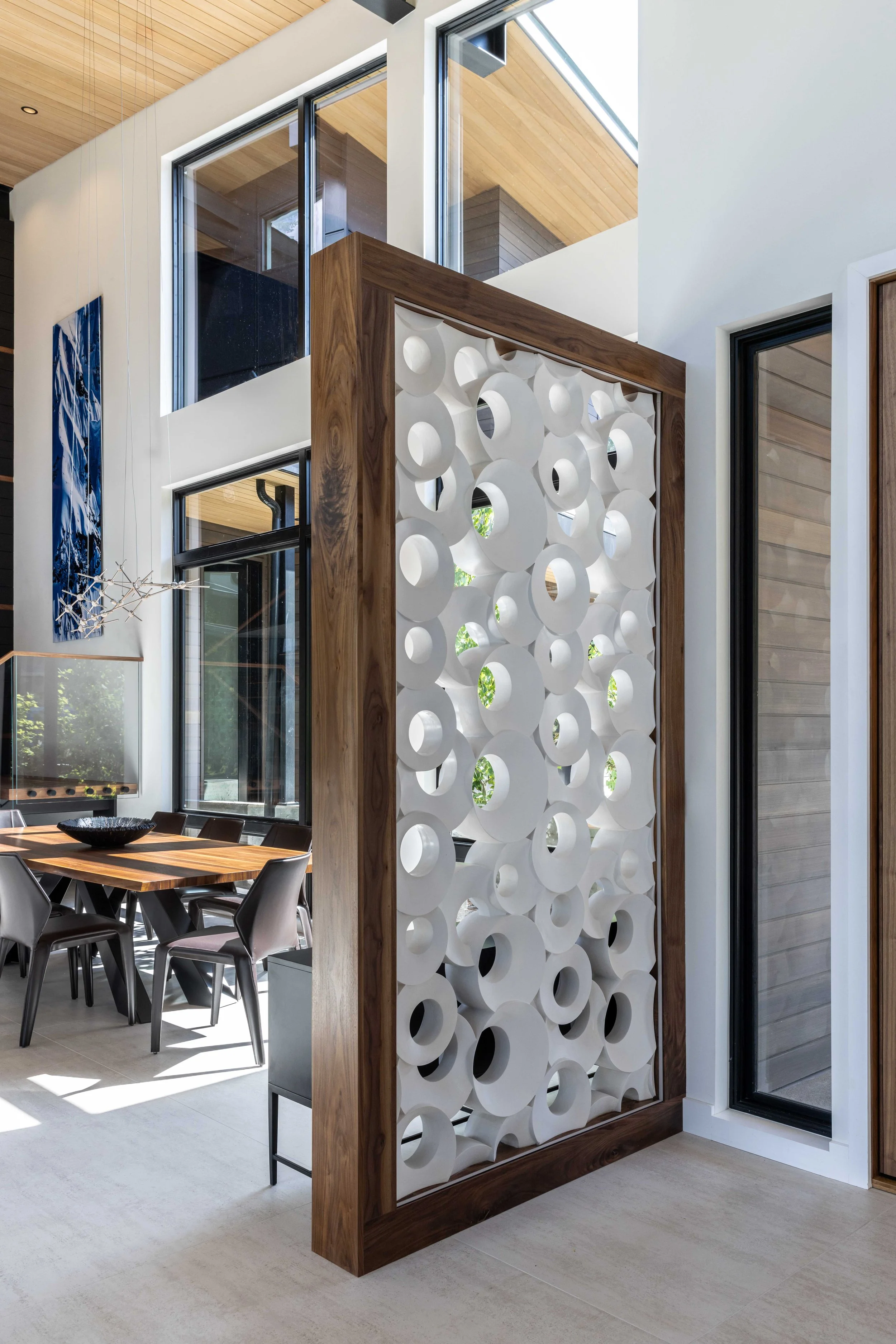 Interior view of a modern house with large windows, a wooden dining table, black chairs, and a decorative white partition with circular patterns.