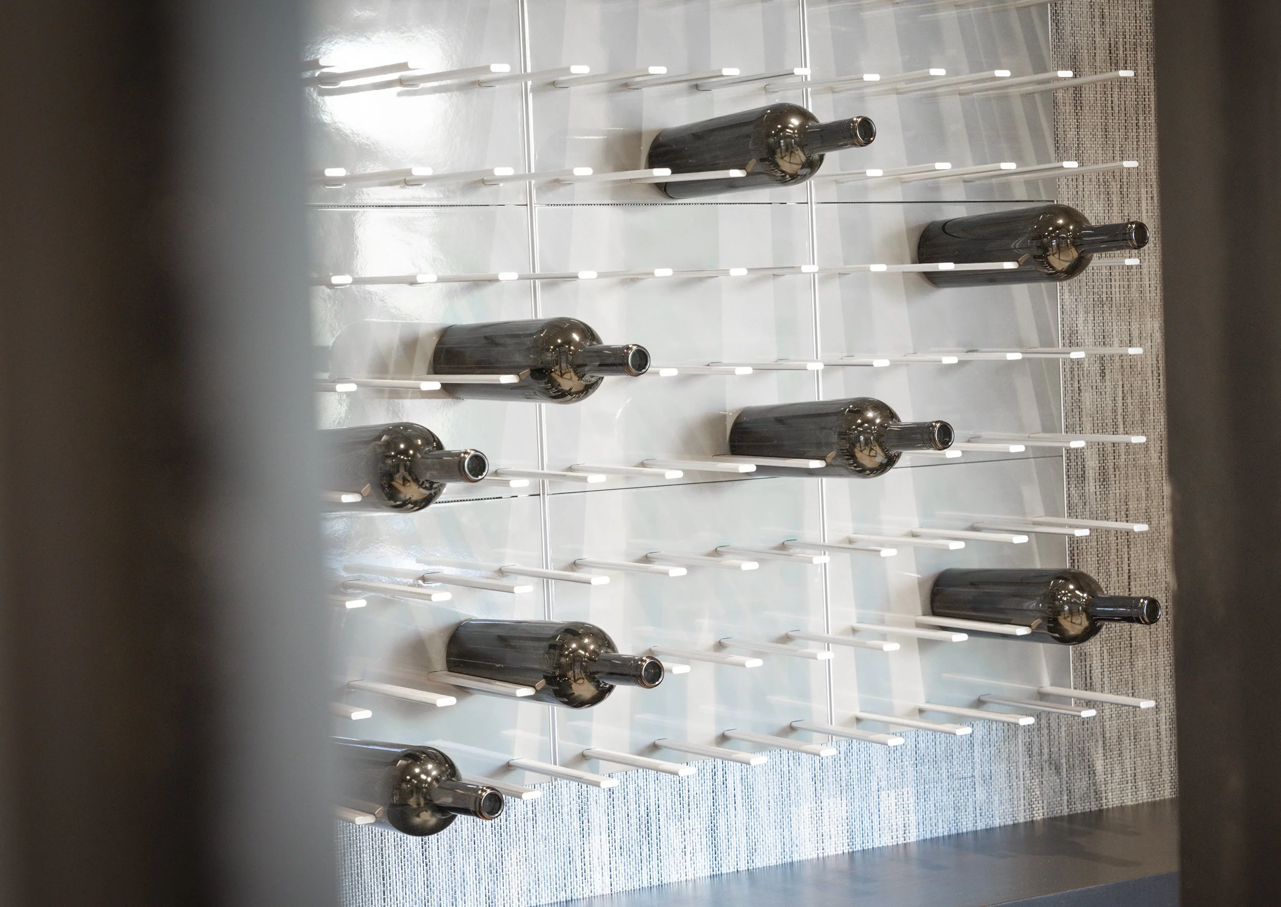 Multiple black glass bottles with long necks resting on a white grid shelf against a white wall, seen through black framing elements in the foreground.