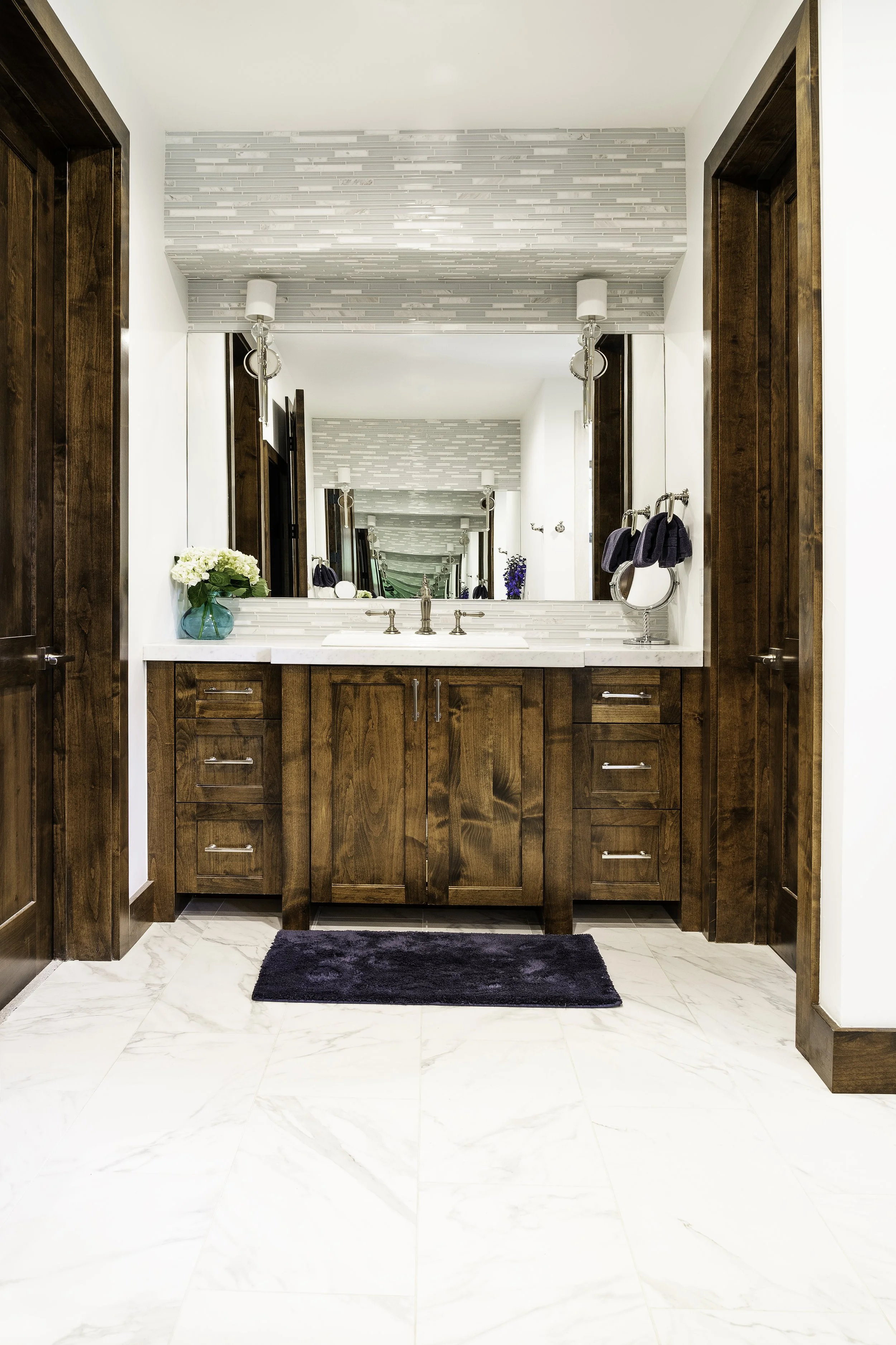 A bathroom with a wooden vanity, marble countertop, large mirror, and white marble-like floor tiles. There are small drawers on each side of the vanity, a black rug in front, and white walls with gray textured accents. Blue towels are hanging on a to