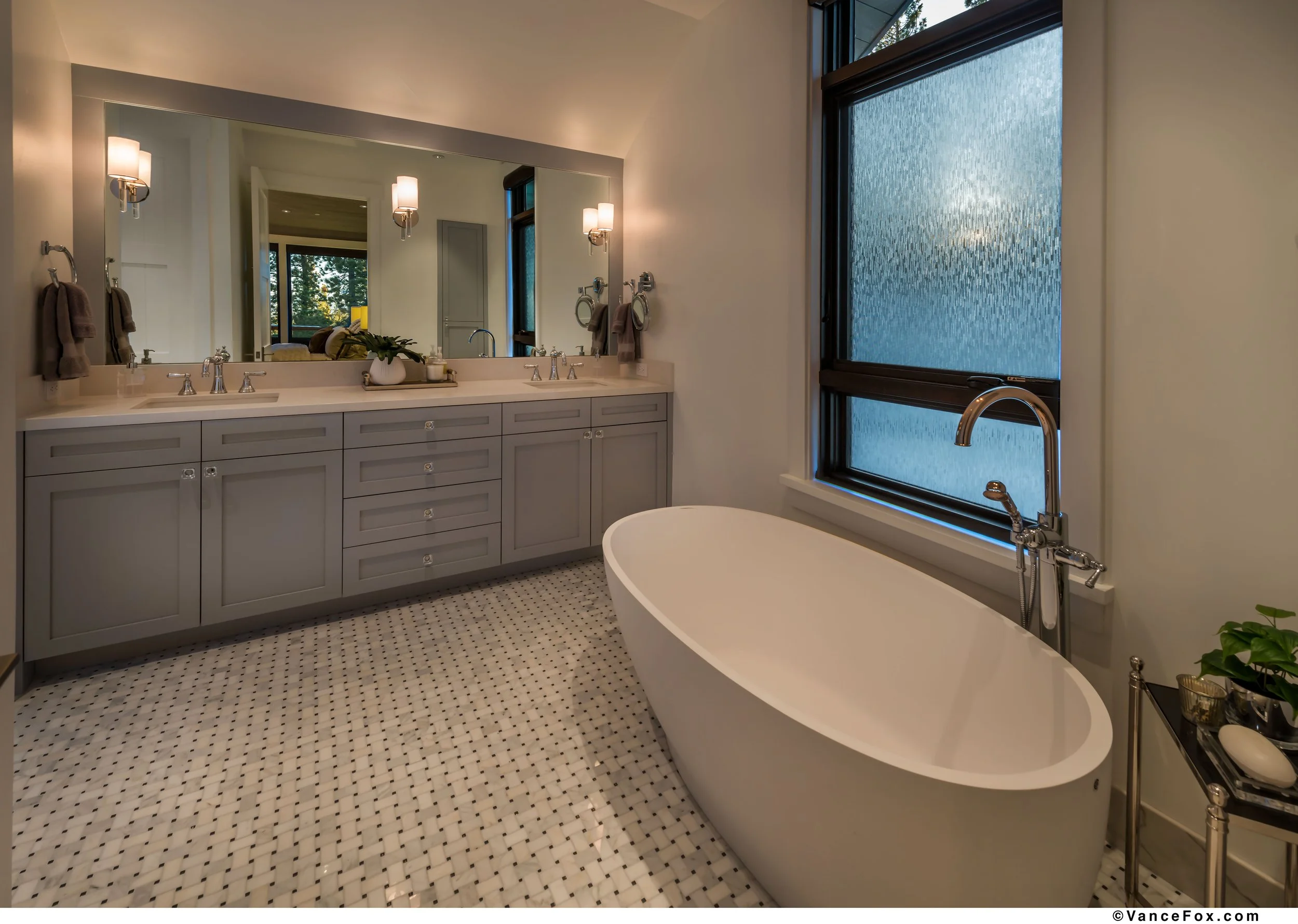 Traditional marble bathroom with a large freestanding soaking bathtub, double vanity with two sinks, large mirror, wall-mounted lights, rain glass window, and a small side table with decor and a plant.