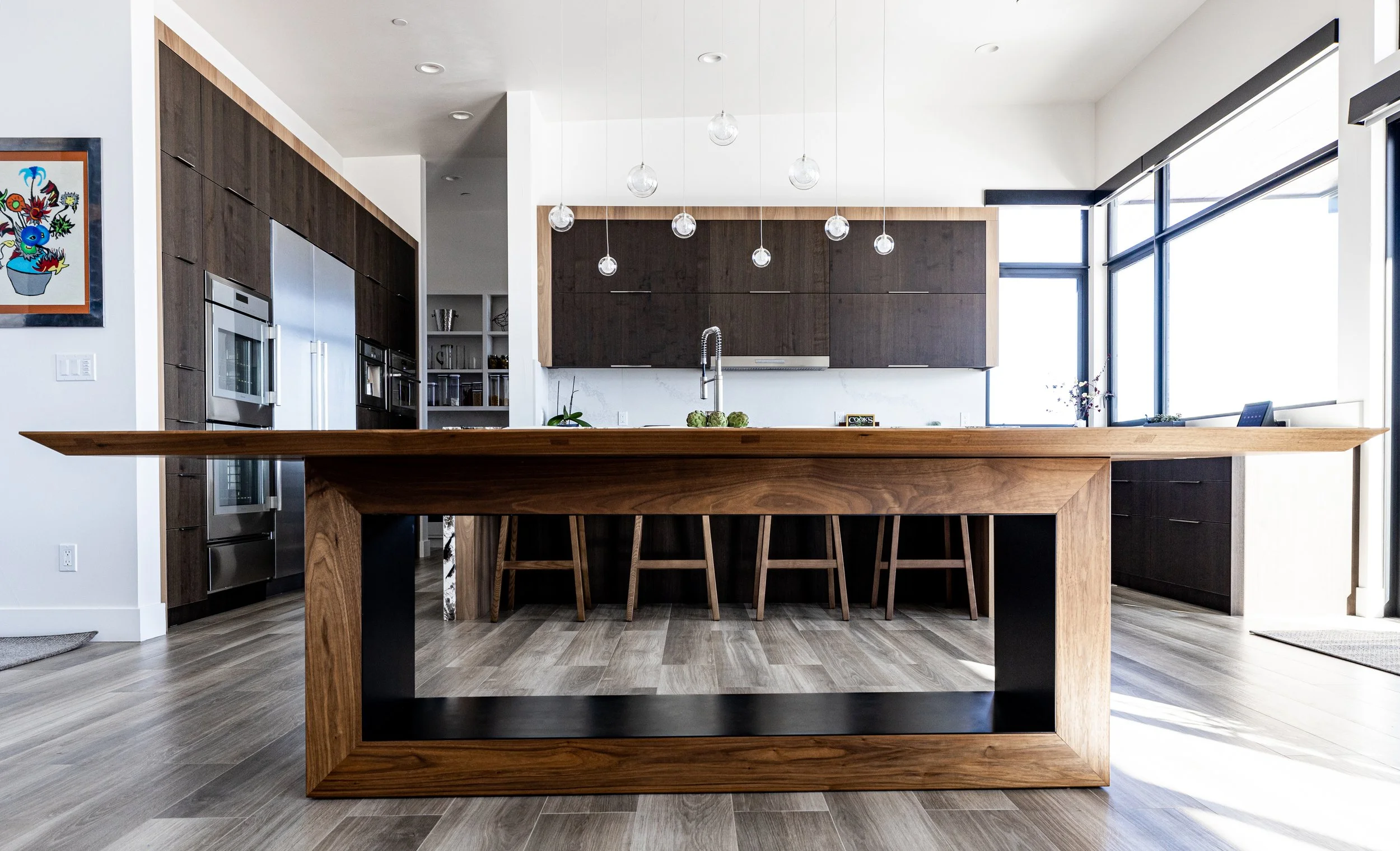 Modern kitchen with a large wooden island, dark wood cabinets, and a row of hanging globe pendant lights, large windows on the right allowing natural light.