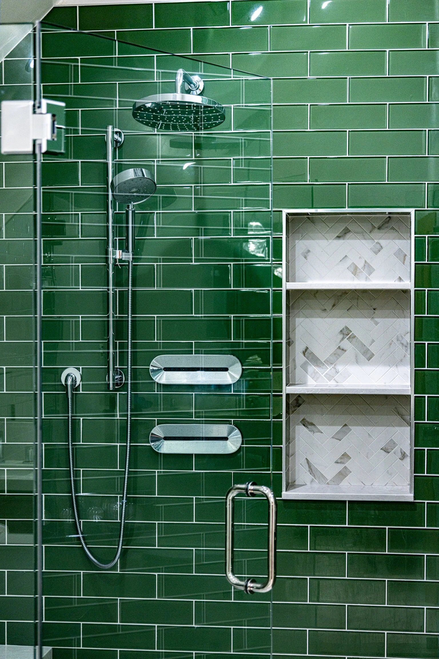 Close-up of a modern shower with green subway tiles, a glass door with a metal handle, two built-in shelves with white herringbone tile, and stainless steel fixtures including a ceiling rain shower and handheld spray.