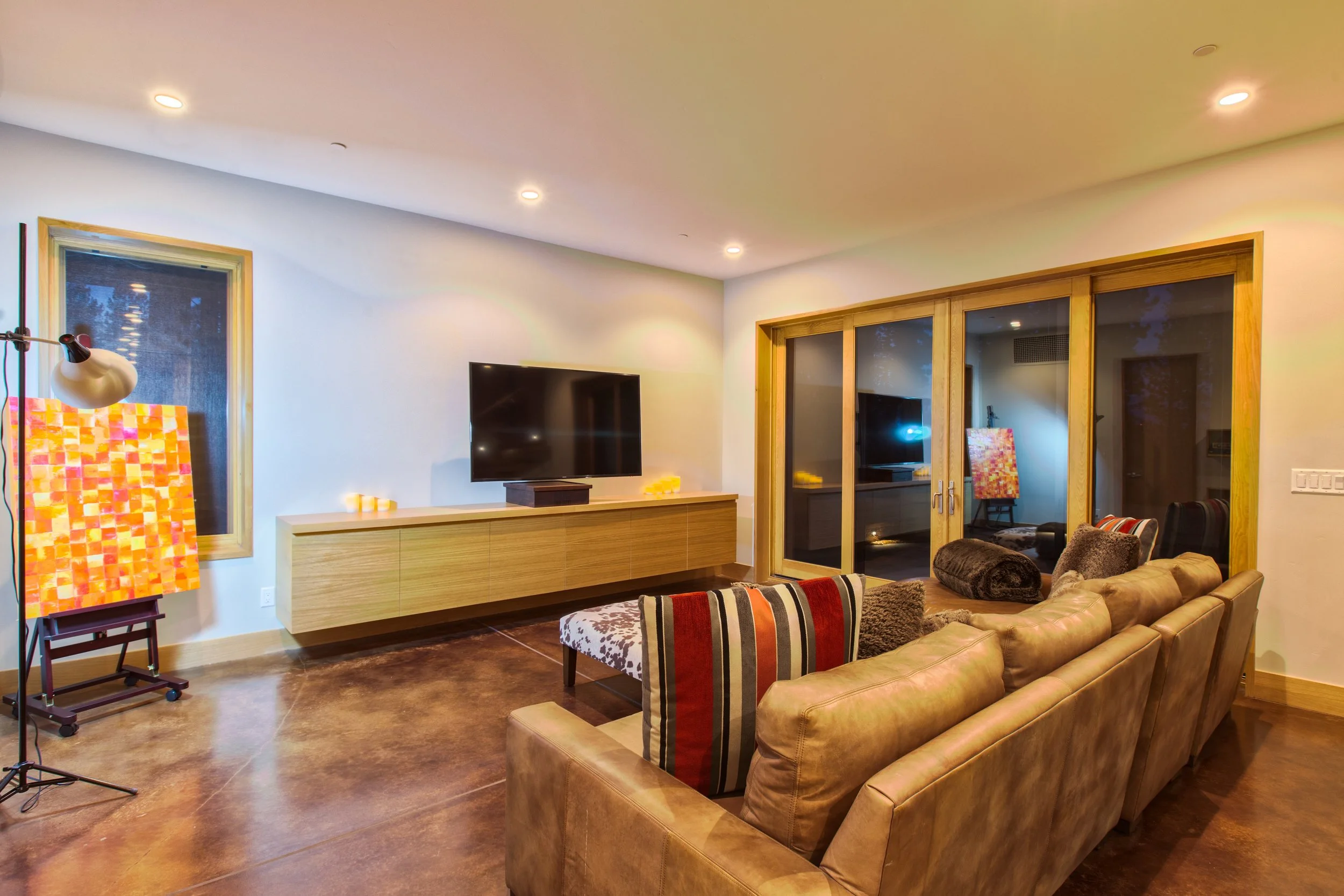 Modern living room with a large brown leather sofa, striped and solid cushions, a wooden TV console with a TV, sliding glass doors, an arched window, and an easel with colorful artwork.