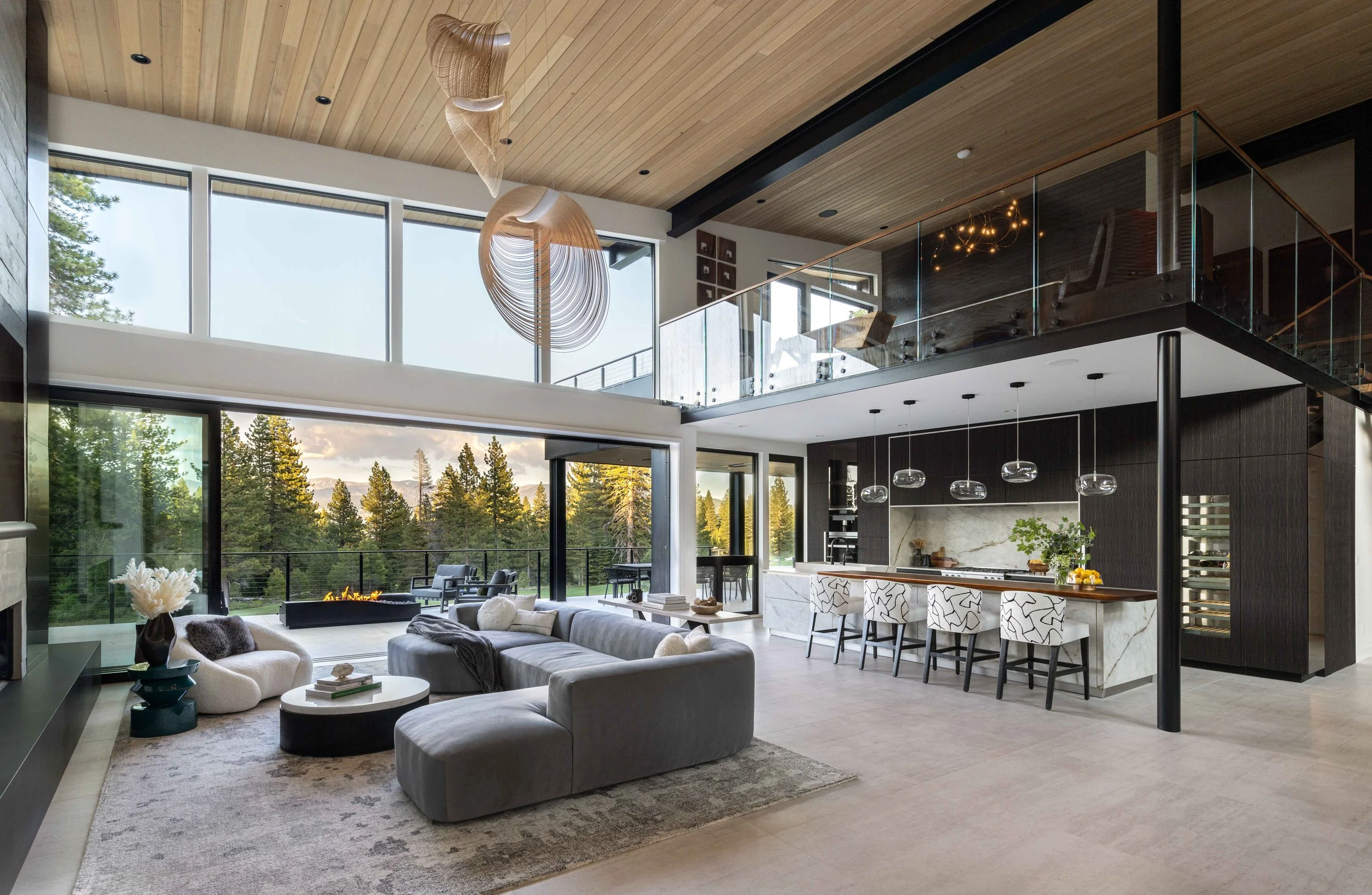Open-concept living room and kitchen with large windows overlooking trees, modern furniture, and high ceiling with wood paneling and hanging artistic light fixtures.
