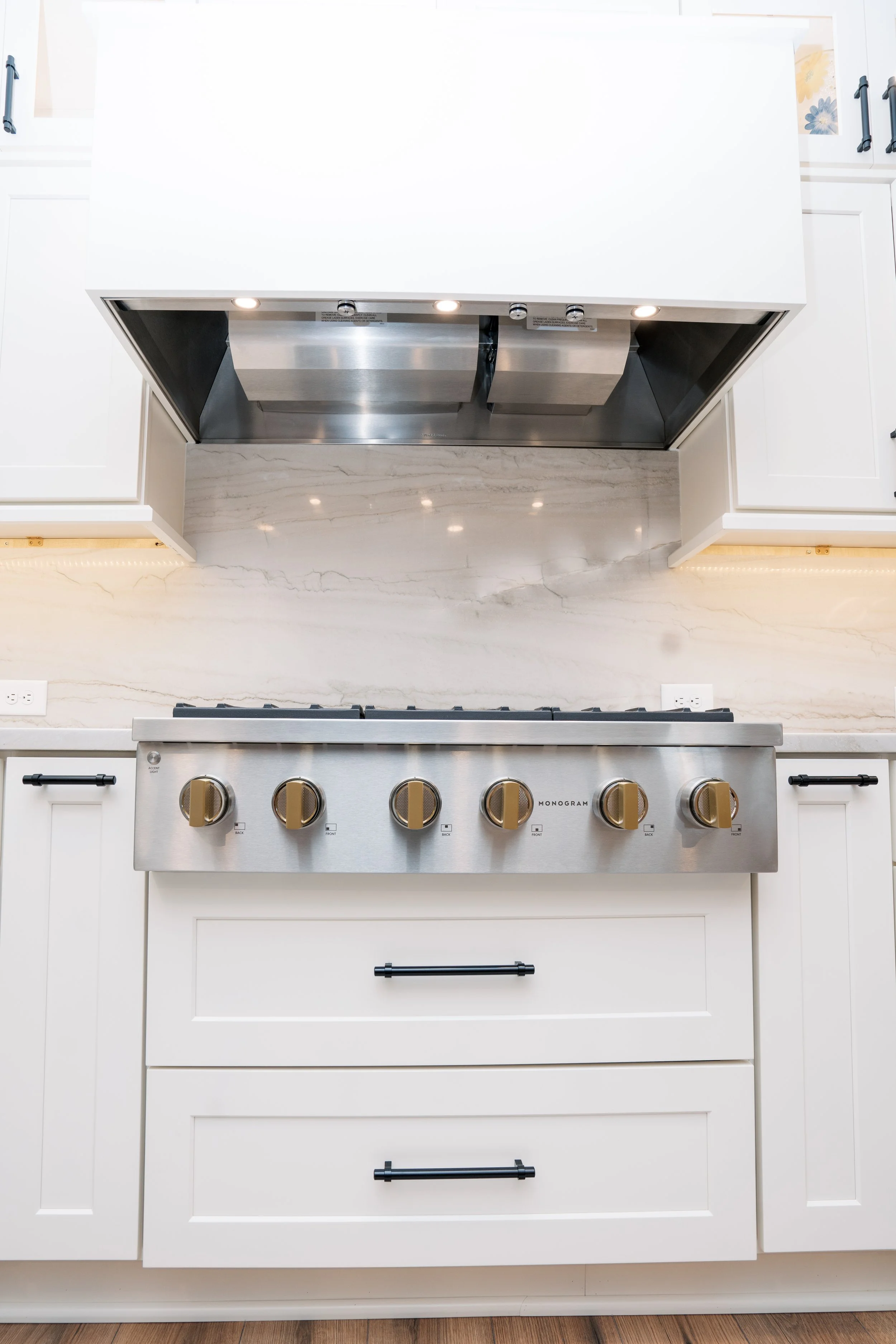Stainless steel gas cooktop with five knobs on a white kitchen cabinet, with a marble backsplash and a range hood above.