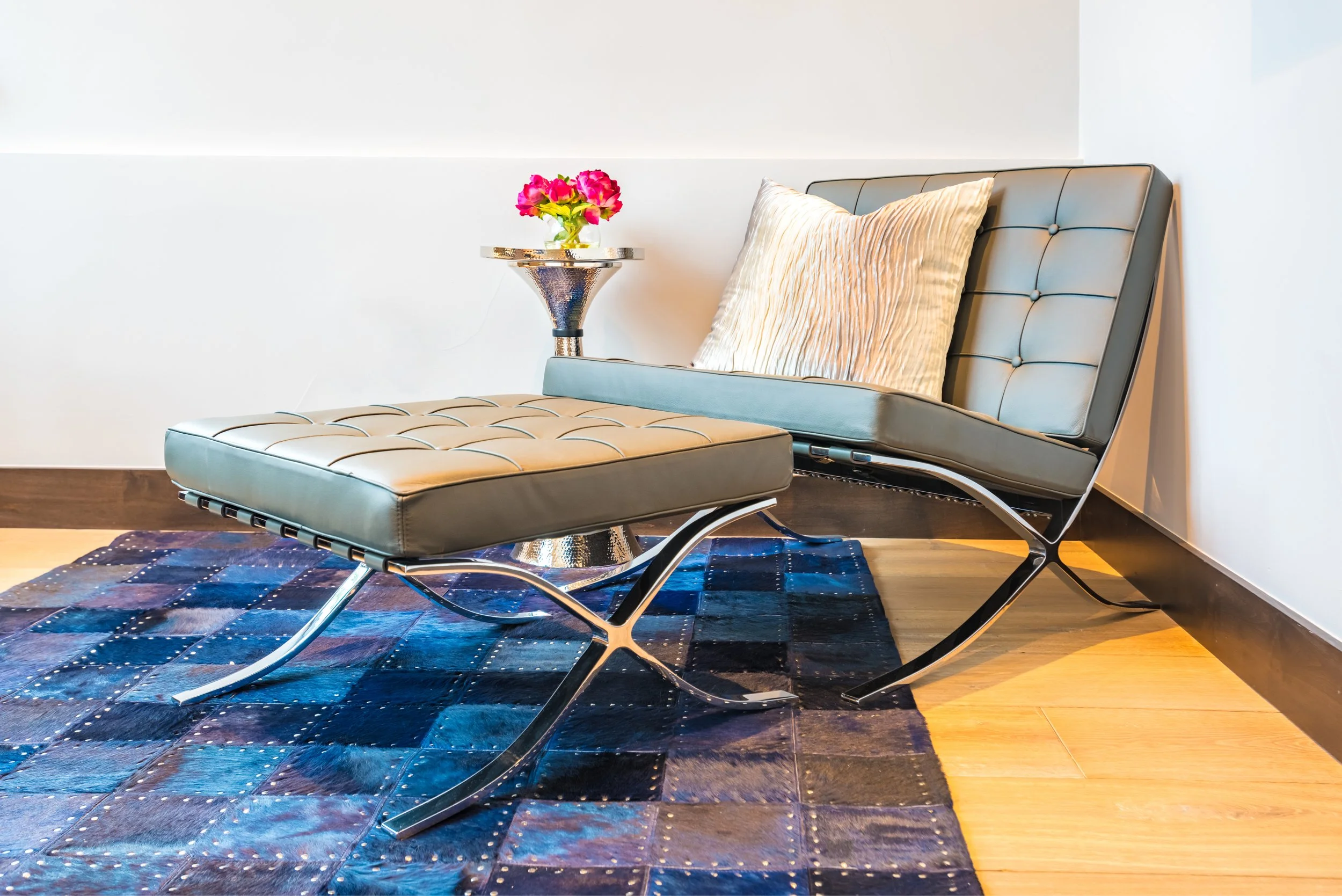 Modern lounge chair with matching ottoman, cream cushion, and a metallic side table with pink flowers, on a blue patterned rug in a well-lit room.