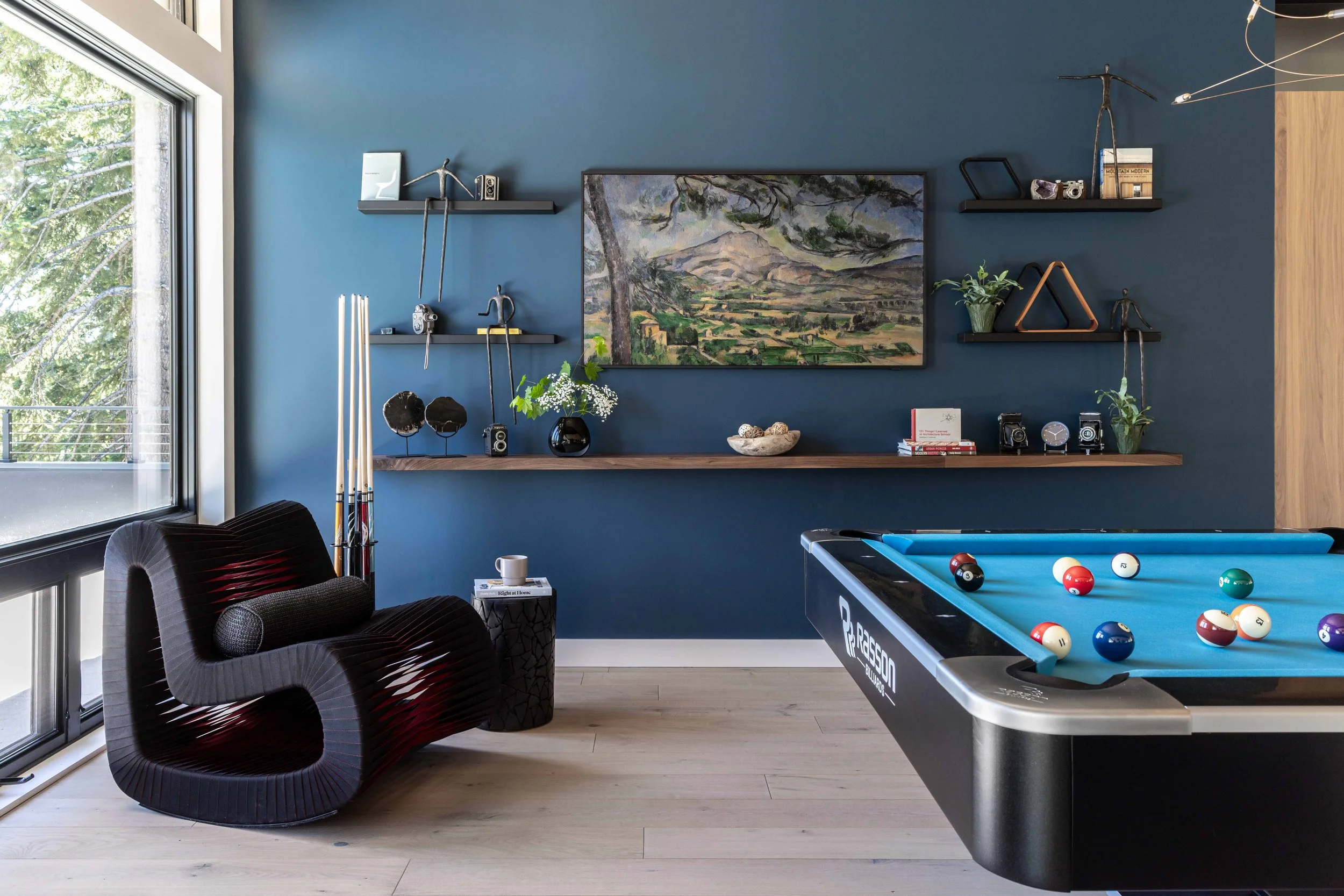 A modern game room with a blue accent wall, a pool table with balls, a black contemporary chair, and floating shelves with decorative items, a landscape painting, and plants.