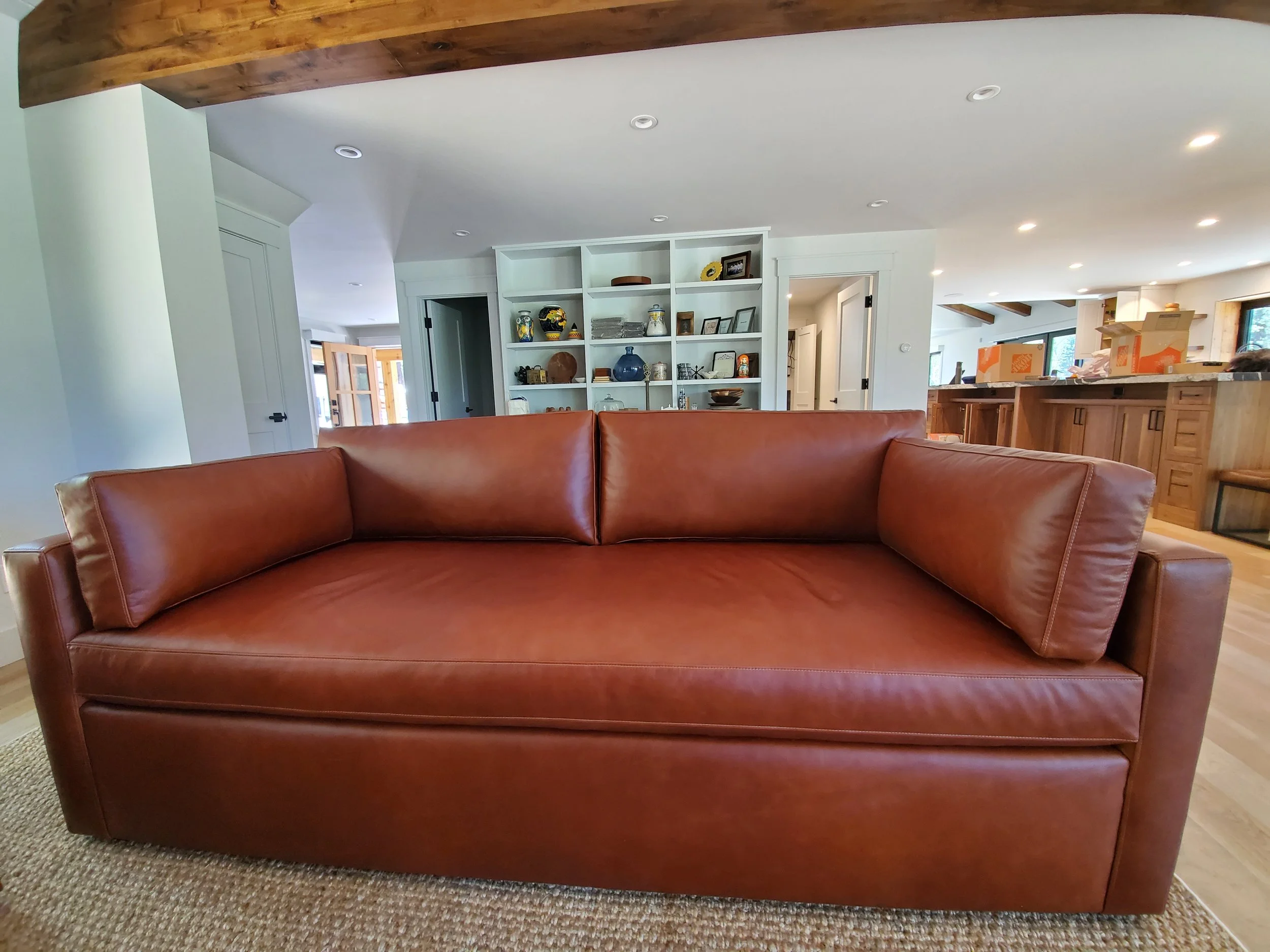 A brown leather couch in a modern, open-concept living space with white walls, wooden accents, and built-in shelves filled with various decorative items and pictures.