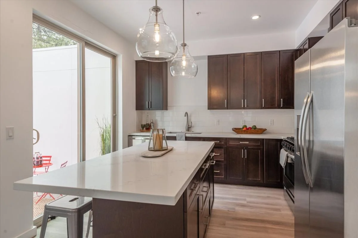 Modern kitchen with dark wood cabinets, white countertops, and stainless steel appliances. A large sliding glass door leading outside, hanging pendant lights, and a wooden bowl with vegetables on the counter.