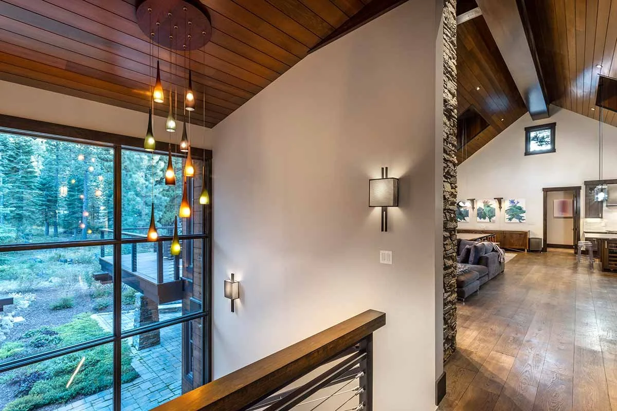 Living room with wooden vaulted ceiling, large window with forest view, modern pendant lights, and wall-mounted lights.