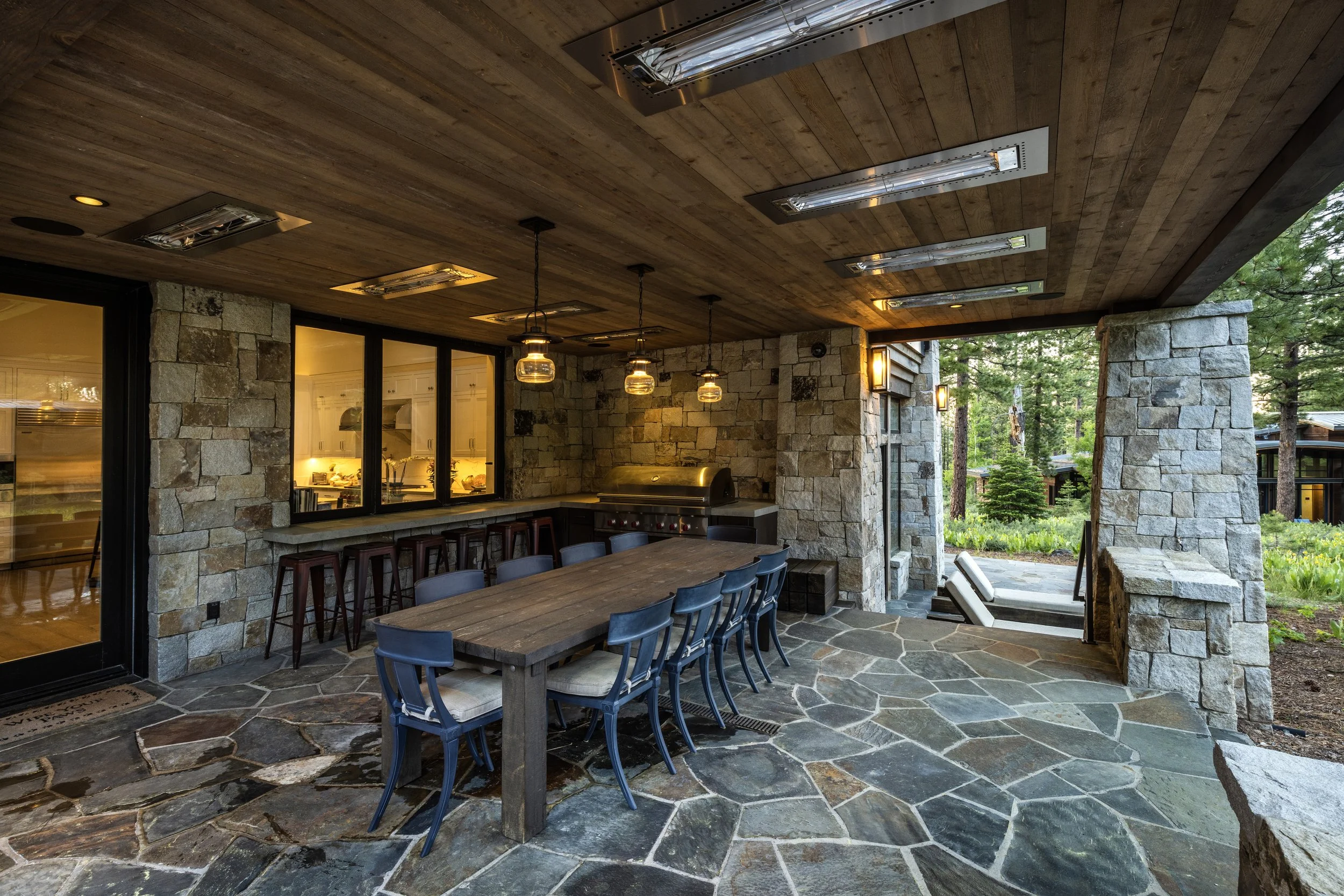 Outdoor covered patio with wooden ceiling, stone walls, and stone flooring. There is a long wooden dining table with blue chairs, a bar area with stools, a built-in grill, and large windows looking into the house. The patio opens to a wooded outdoor 