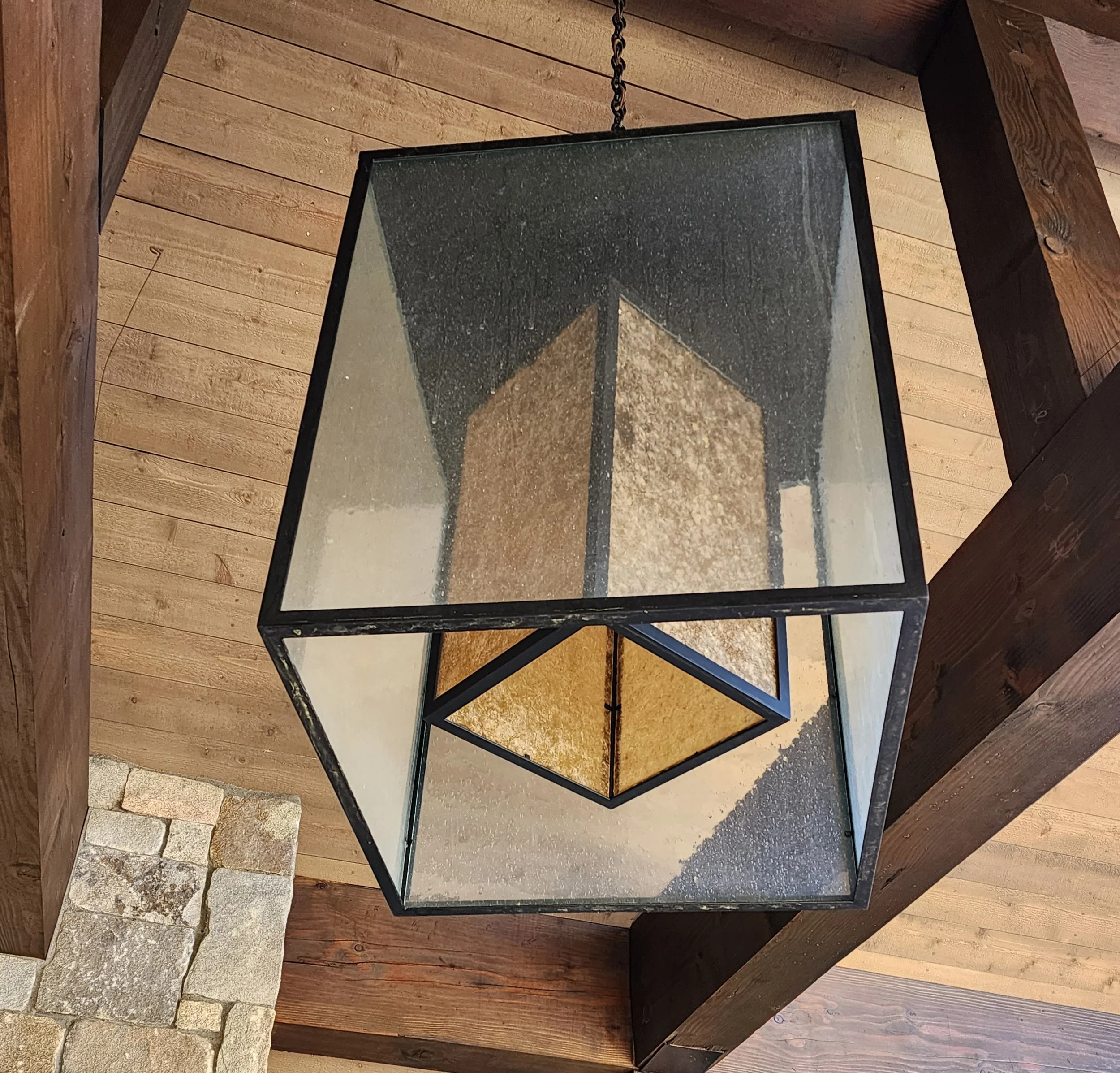 A modern geometric pendant light fixture hanging from a wooden ceiling, with a combination of black metal frame and glass panels on the outside and a mica and black framed interior shade. Custom designed by Emily Roose.