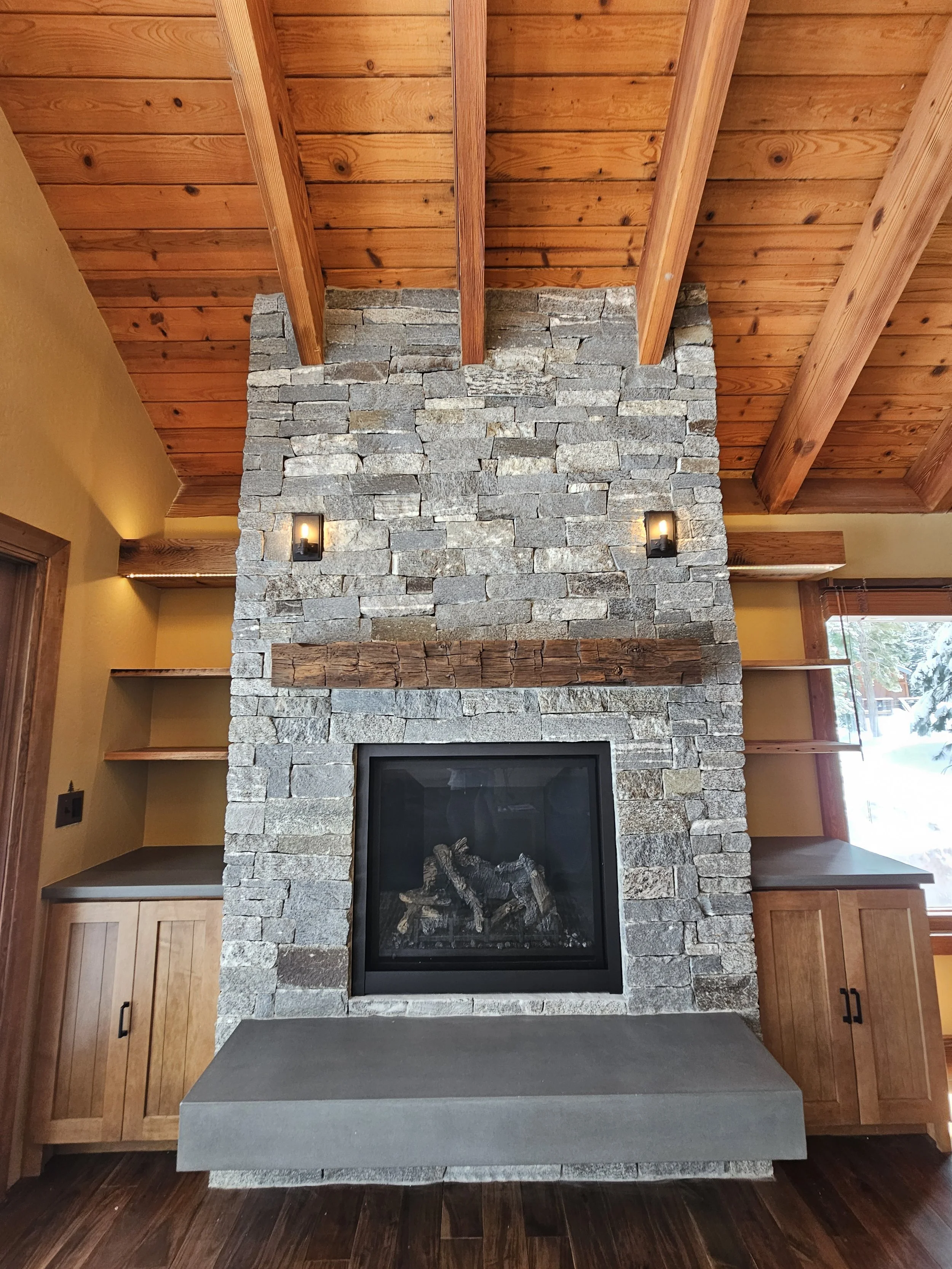 Interior view of a living room featuring a large stone fireplace with a wooden mantel, flanked by built-in shelves and cabinets. The ceiling has exposed wooden beams, and there is a window on the right side showing outdoor snow.