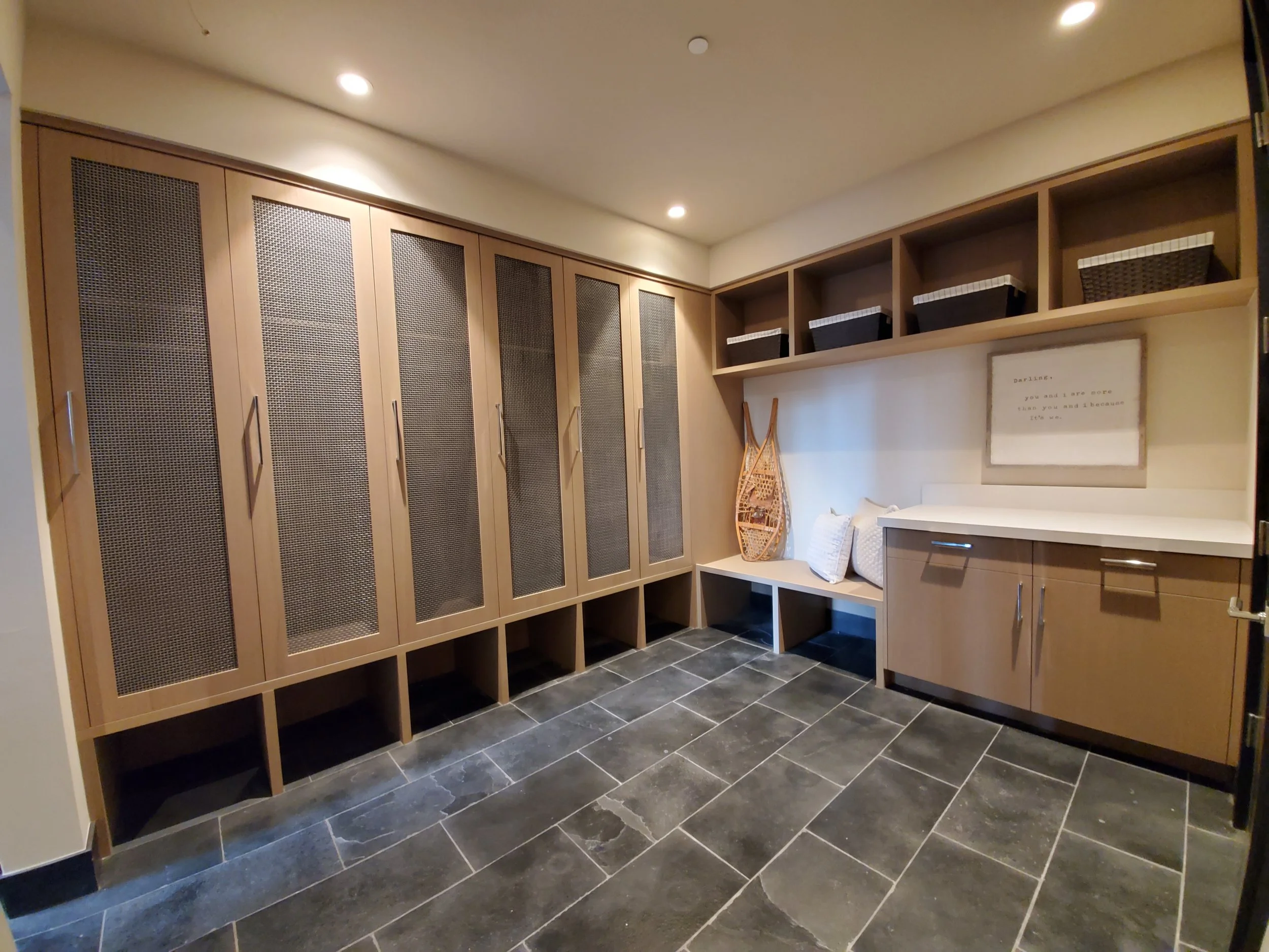 A well-organized room with wooden cabinets, open shelving, and a bench with decorative pillows. Baskets are stored on the upper shelves and a framed quote hangs on the wall. The room has a dark tile floor and ceiling recessed lighting.
