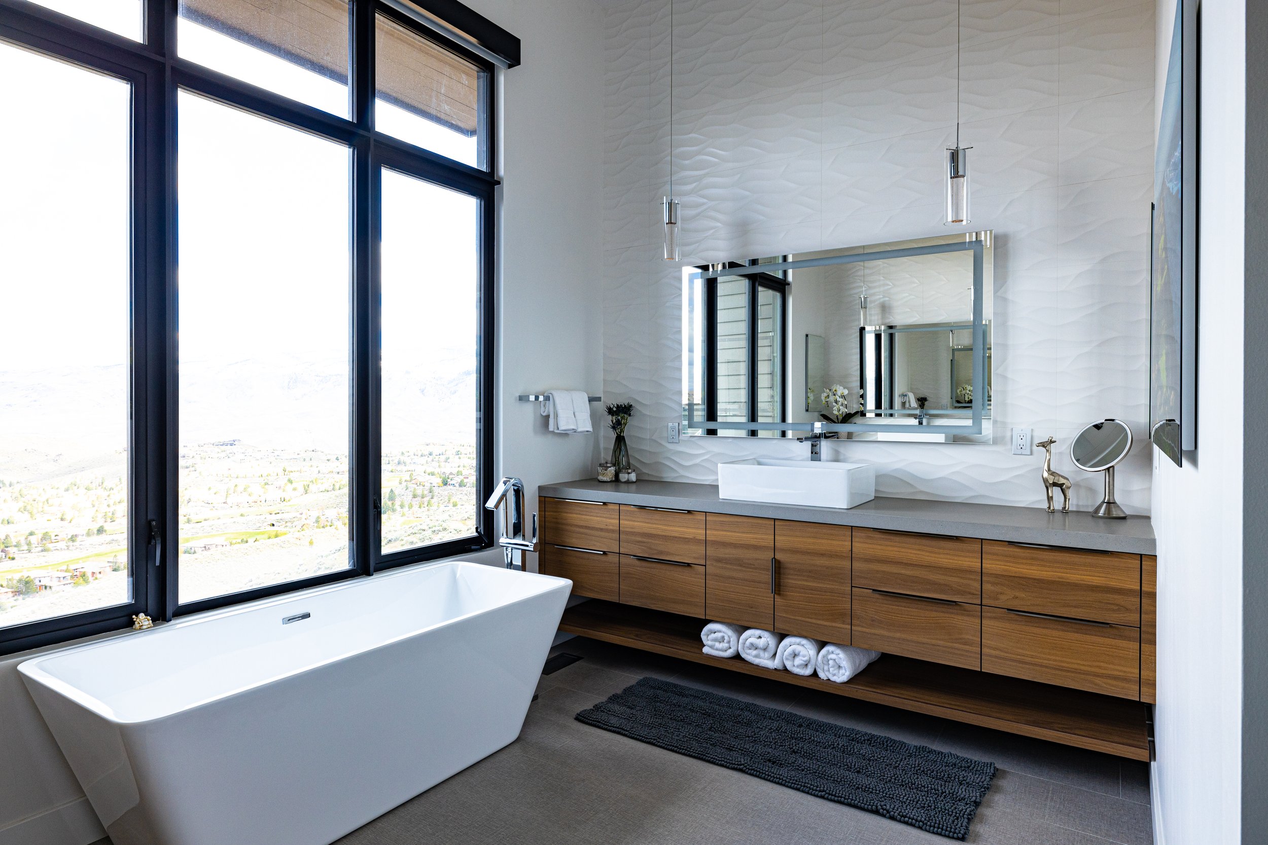 Modern bathroom with a large window, white bathtub, wooden vanity with six drawers, a rectangular white sink, silver faucet, large mirror, small decorative items, and a dark rug, overlooking a landscape.